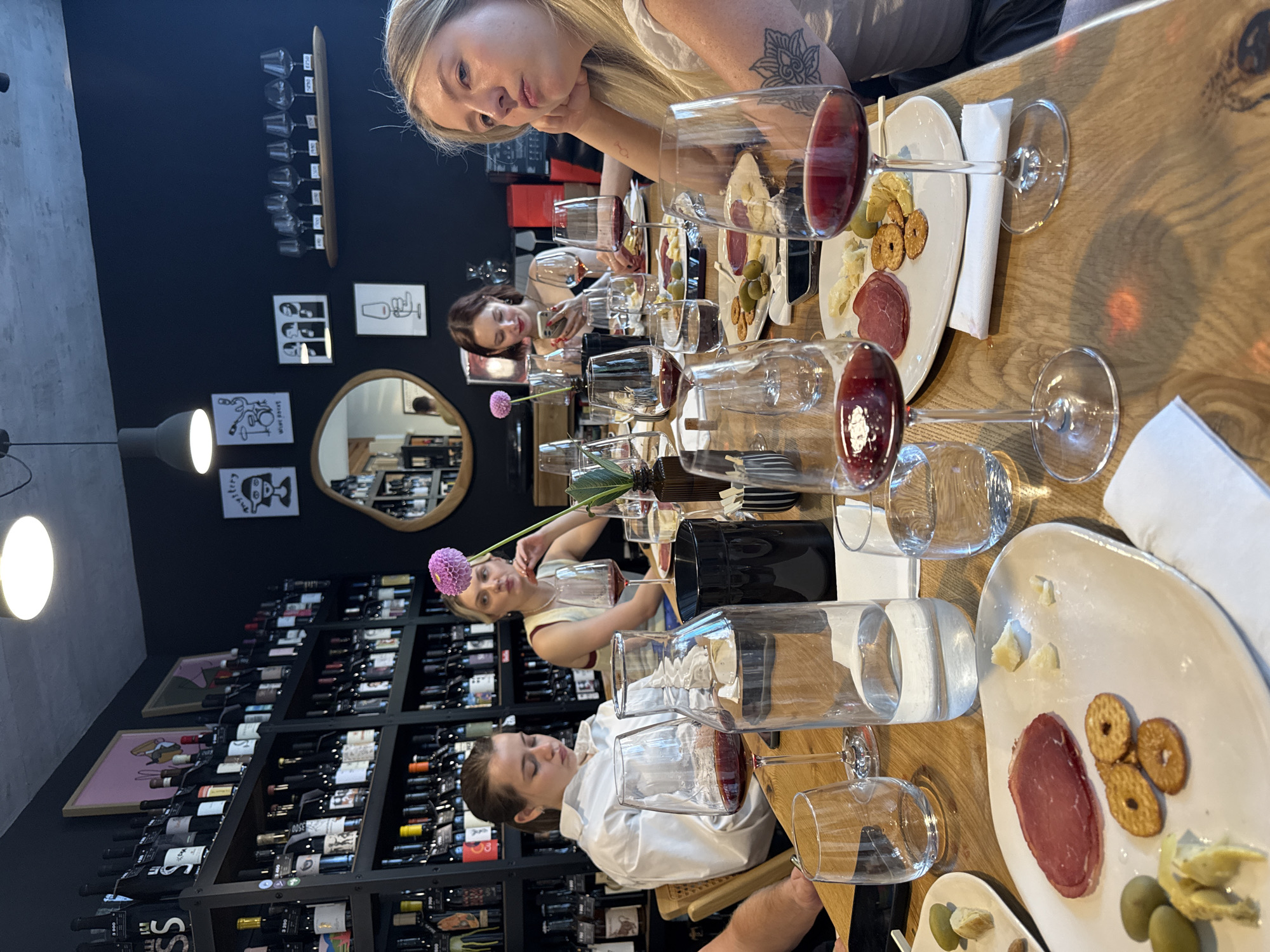 An indoor, slightly elevated, eye-level shot captures a group of individuals, primarily young women, seated around a light-colored wooden table at what appears to be a wine bar or restaurant, described as being in Vracar Urban Municipality, Serbia. The scene is dominated by the long wooden table, laden with multiple wine glasses, plates of food, and other serving ware. Several stemmed wine glasses, many containing red wine, are prominently displayed across the table. White ceramic plates hold various small food items including slices of cured meat, green olives, small crackers or pretzel-like snacks, and chunks of cheese. A clear glass carafe filled with water, along with smaller tumblers, are also present. White paper napkins are scattered on the table. A dark-colored cylindrical container, possibly holding cutlery or another item, is visible. A single purple dahlia flower is seen near one of the seated individuals. Five individuals are visible at the table. In the immediate foreground, to the right, a young woman with long blonde hair, wearing a light-colored top, rests her chin on her left hand, gazing towards the right of the frame with an introspective expression. A floral tattoo is visible on her left forearm. To her left, further down the table, a young woman with dark hair, clad in a white long-sleeved shirt, is looking downwards, seemingly at a mobile phone held in her hands. Behind her, another young woman with blonde hair, wearing a light-colored sleeveless top, is visible, looking slightly to her left. Her right hand is near her chin. Further in the background, to the right, another young woman with dark hair, wearing a dark top, is also observed looking at a mobile phone. A fifth individual, partially obscured, is visible on the far left side of the table, only their arm and shoulder are discernible. The background features dark, possibly black or very dark blue, walls. Extensive black shelving units, densely packed with numerous wine bottles of various shapes and labels, line the left and rear walls, indicating a specialized wine establishment. The ceiling appears to be concrete with suspended pendant lights. Several framed artworks and decorative items adorn the walls: on the left, an abstract piece with a pink background; on the right, multiple black-and-white minimalist prints depicting figures and objects (e.g., a person with a 'mystery' thought bubble, a drum set, multiple faces). An oval-shaped mirror with a wooden frame is mounted