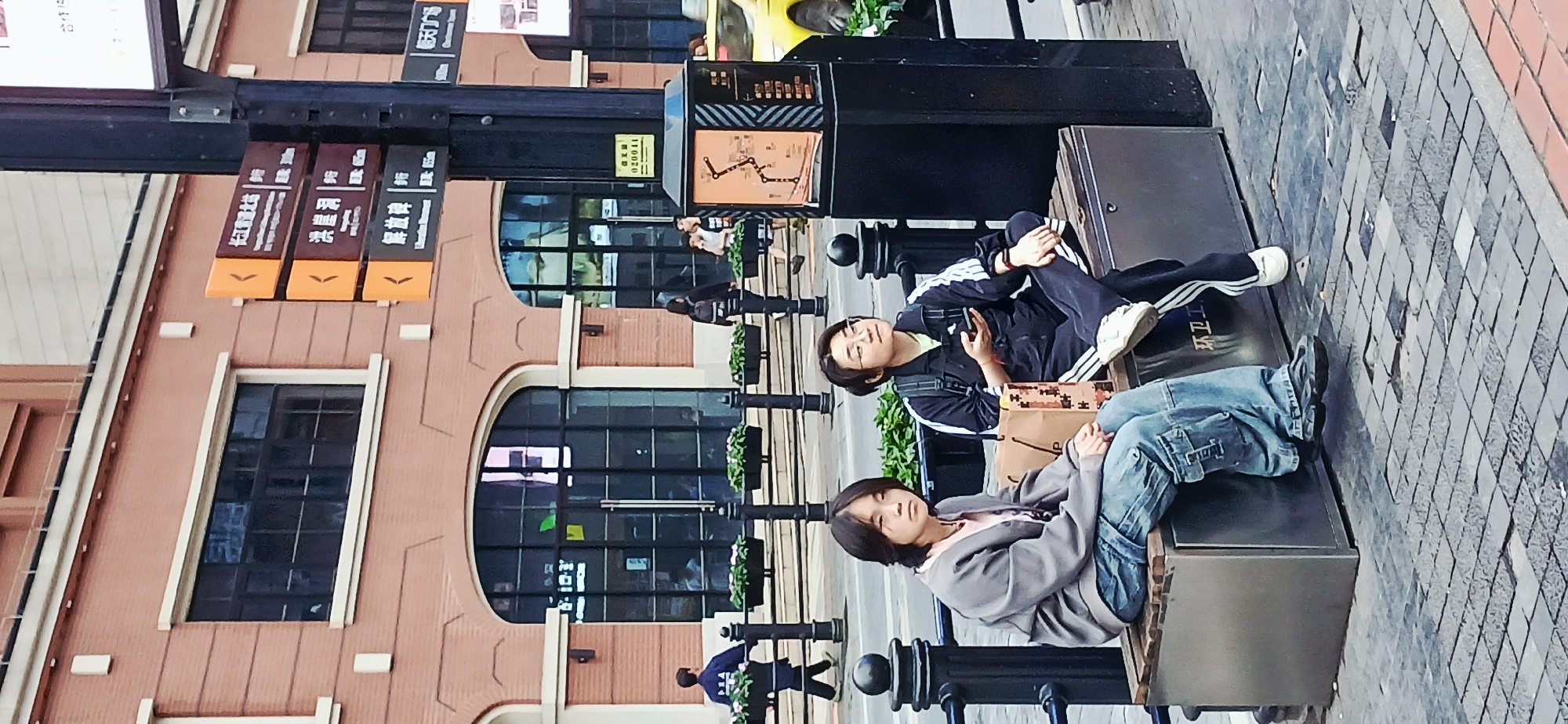 Two people, both appearing to be young adults, are reclining on a bench in an outdoor urban setting. The first individual, positioned at the top of the frame, is wearing a dark tracksuit with white stripes, white sneakers, and is holding a cell phone. A backpack and shopping bag are also visible. The second person is wearing a gray sweater and blue jeans, and has dark hair. They are both lying on a metal bench with cast iron uprights, possibly located on a city sidewalk. In the background, there is a brick building with arched windows and street signs with Chinese writing. Other pedestrians are visible walking in the distance. The location is Yuzhong District, China.