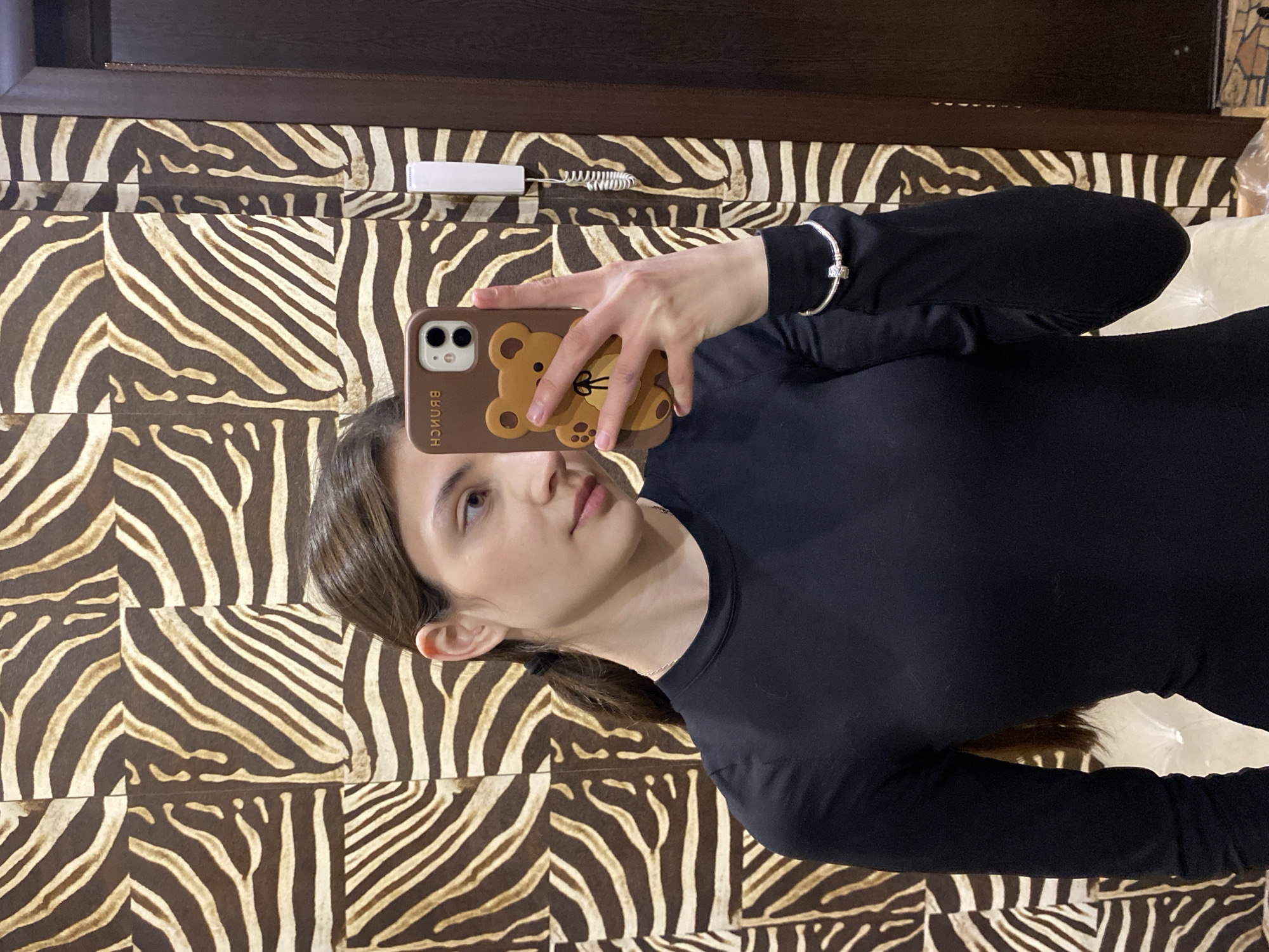 A woman is taking a selfie. She is wearing a black, long-sleeved shirt and a silver bracelet on her left wrist. She is holding a smartphone in a brown case with a teddy bear design. The word "Brunch" is written vertically on the case. The background is a wall covered in brown and white zebra-striped tiles. A cleaning brush with a white handle is visible on the wall above her head. The location is unknown and not specified.