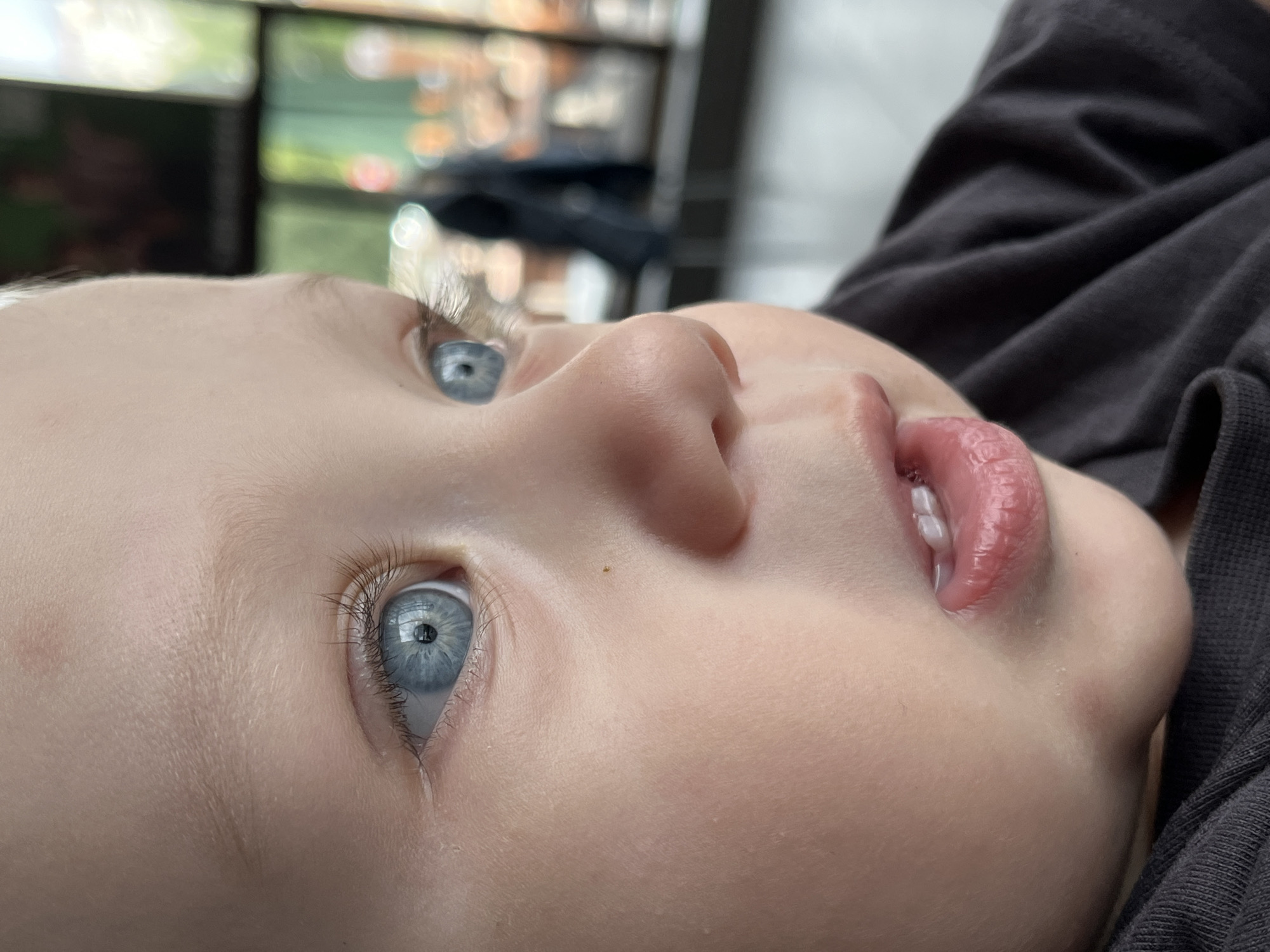 A close-up, side-profile photograph captures a young child's face, focusing on their striking blue eyes. The child's skin is fair and smooth, with a hint of pink across their cheeks and chin. Their lips are slightly parted, revealing two small teeth. The background is blurred, suggesting an indoor setting with some daylight filtering in, possibly a cafe or restaurant, with patrons and tables faintly visible. The image is well-lit, highlighting the details of the child's features. There is no information in the image to suggest a location in Nizhny Novgorod, Russia.