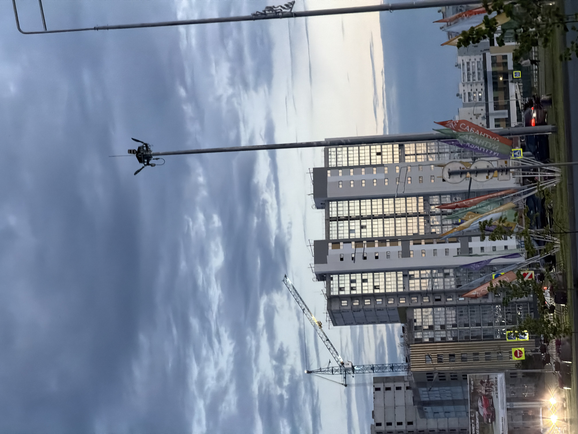 This is a vertically oriented photograph taken outdoors, likely in Nizhnekamsk, Russia. The scene captures an urban environment with multiple high-rise buildings, some appearing to be under construction or recently completed. A large construction crane is visible, with its arm extended towards the sky, suggesting ongoing building activity.
The sky is overcast with dramatic, textured clouds, hinting at a potentially unsettled or approaching storm, or perhaps the aftermath of one. Despite the cloudy conditions, there's a bright glow in the upper right portion of the sky, indicating either late afternoon or early morning light breaking through.
Several colorful flags or banners are strung along wires or poles, some bearing Cyrillic text. One banner clearly reads "САБАНТУЙ" (Sabantuy), which is a traditional Tatar festival. Another banner shows "САБАНТУЙ" as well. This suggests the photograph might have been taken around the time of this festival.
In the foreground, a tall pole extends upwards, with a device resembling a camera or sensor attached to it near the top. This could be part of a surveillance system, a weather monitoring station, or broadcasting equipment.
Roads and traffic are visible in the background, with cars present, including one with its brake lights illuminated. Street signs, including pedestrian crossing signs with yellow backgrounds and red circles, are also noticeable. The overall impression is a dynamic urban landscape undergoing development, with cultural elements (the banners) adding a specific context. The lighting suggests it's either dawn or dusk.