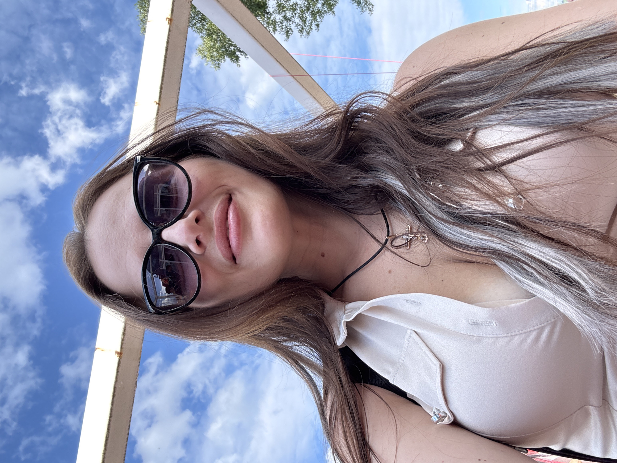 The media file features a close-up, low-angle selfie of a woman outdoors. The woman has long, dark hair with streaks of gray and is wearing large, dark sunglasses. She is also wearing a light-colored, button-up shirt. A delicate, cross-shaped necklace is visible around her neck. The background shows a bright blue sky with scattered white clouds, suggesting it is daytime with pleasant weather. There is also a hint of some outdoor structure, possibly a pergola or canopy, with beige-colored beams and some greenery, indicating a park or garden setting. The image is a self-portrait, likely taken during a casual outing.