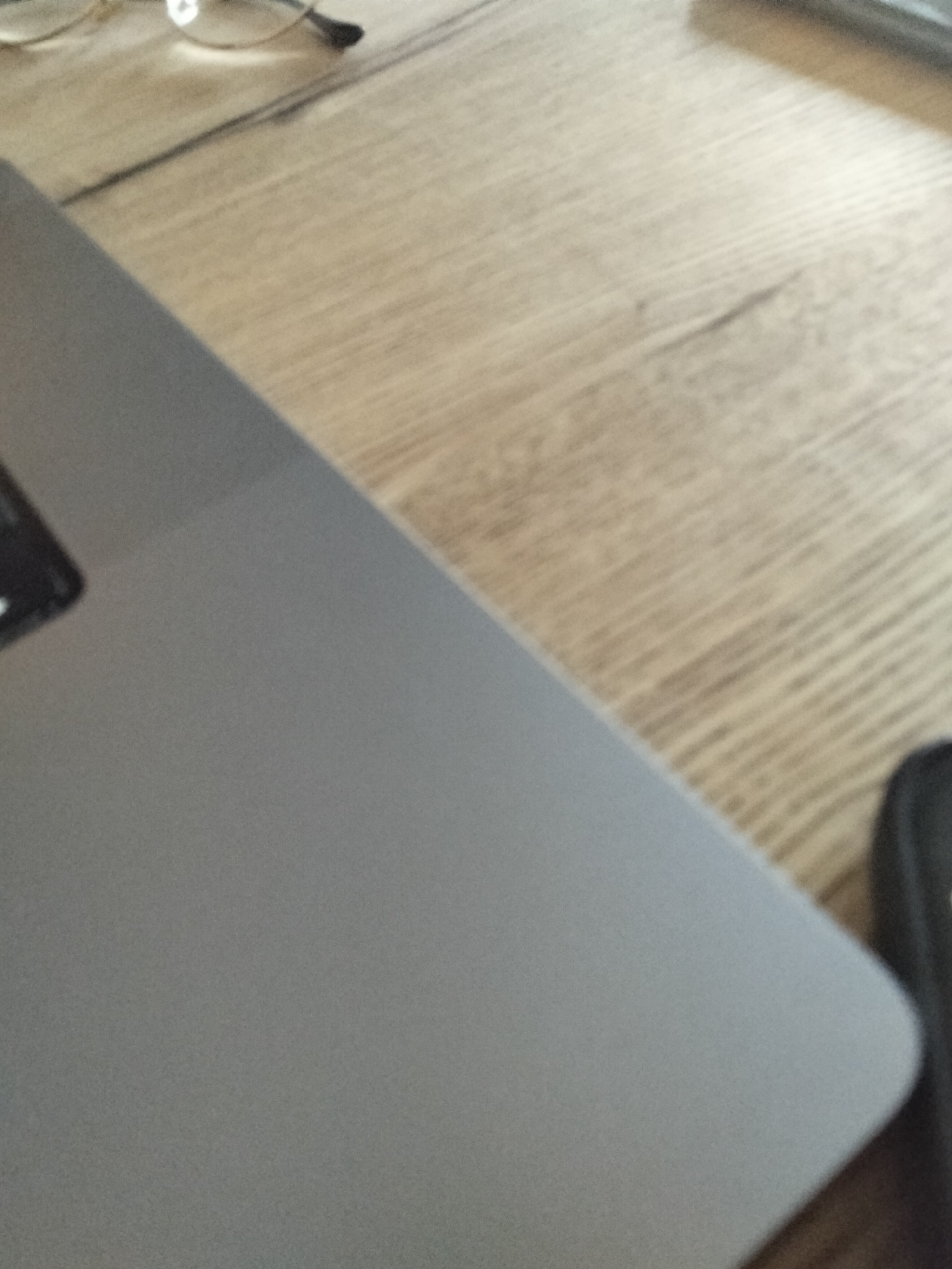 The image shows a close-up view of a laptop on a wooden desk. The laptop is silver and takes up a significant portion of the frame, angled diagonally from the bottom left corner towards the top right. Part of the laptop's keyboard, or possibly a trackpad, is visible on the left side. To the right of the laptop, the wooden desk surface is visible with a prominent wood grain pattern. In the upper left corner, out of focus, a pair of eyeglasses with thin gold frames can be seen. In the far right of the image, a dark object, possibly a phone or a case, is partially visible. The lighting appears to be natural, casting soft shadows. The scene is static and appears to be an indoor setting, likely an office or home workspace. There are no people or discernable actions taking place. The location is unknown.