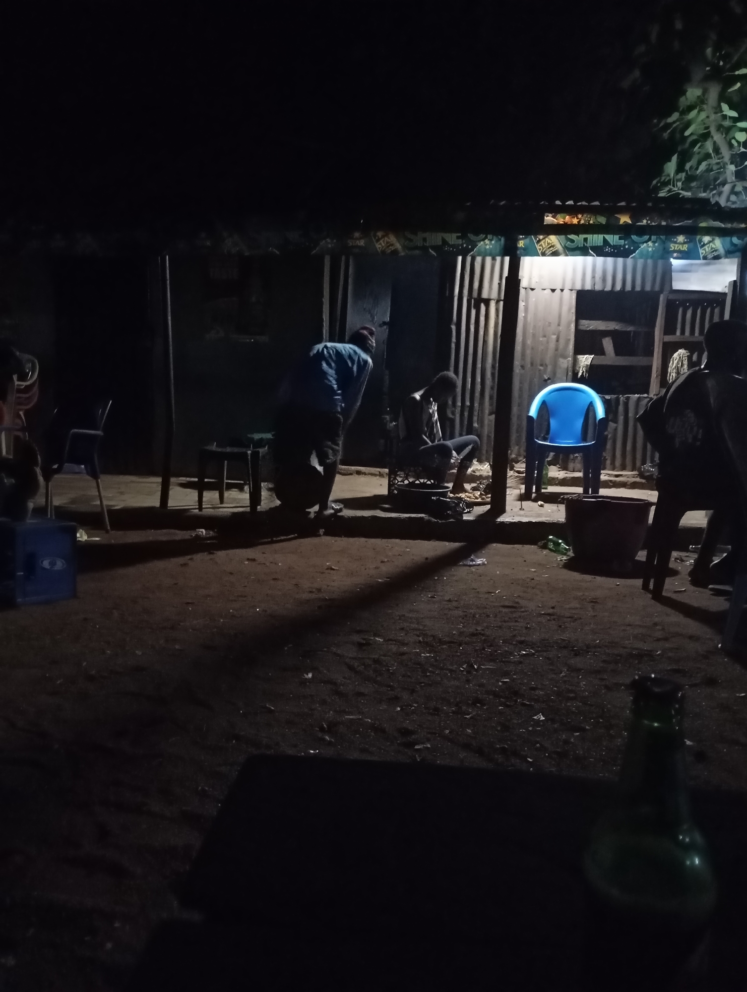 A nighttime scene shows a group of people gathered outside what appears to be a small shop or bar with a corrugated metal exterior. The area is dimly lit, with a single blue chair visible in the foreground. One person is seated at a table, appearing to be preparing food. Another person is bent over, possibly tending to a grill. Several chairs are scattered around the area, and a few bottles are on the ground. The ground is dirt and debris. The shop facade is adorned with banners advertising "STAR" beer and "SHINE". The overall impression is one of a casual, informal gathering in an outdoor setting.