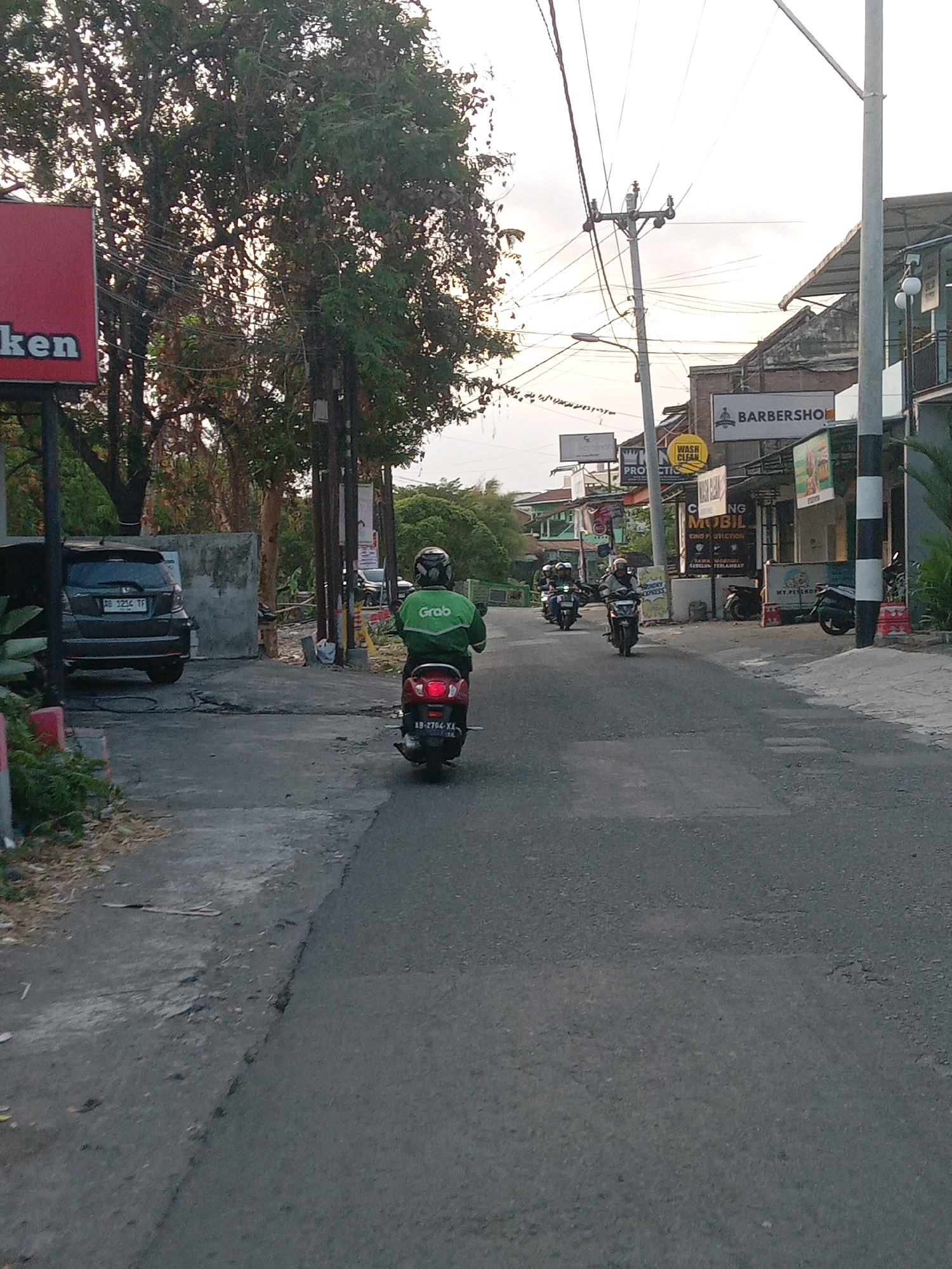 The image captures a street scene in Condongcatur, Indonesia, likely during the late afternoon or early evening due to the soft, diffused light. The perspective is from behind a person riding a scooter, facing forward into the street.
In the foreground, the asphalt road is visible, showing signs of wear with cracks and patches. To the left, a grey hatchback car is parked, with its license plate reading "AB 1234 TF". A red sign with white lettering, partially visible and reading "ken", stands on a pole to the far left. Adjacent to the car, some greenery and a concrete wall are present.
The main subject of the image is a person wearing a green jacket with the "Grab" logo, riding a red scooter. The scooter's license plate reads "AB 2704 XA". The rider is wearing a helmet. Further down the street, several other scooters, also with riders, are seen moving in the same direction.
The environment appears to be a commercial area with various establishments lining the right side of the street. Signs visible include "BARBERSHOP", "WASH CLEAN", and "NG MOBIL KING PROTECTION". Electrical wires are strung across utility poles, a common sight in urban Indonesian settings. Trees with dense foliage are present on the left side of the street, partially obscuring the sky. The overall impression is one of a moderately busy street in a developing urban area.