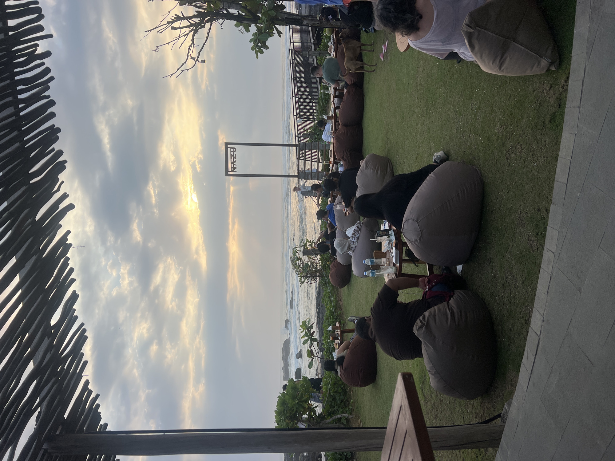 The image depicts a seaside restaurant in Mengwi, Indonesia, during a cloudy sunset. The foreground is occupied by beanbag chairs, tables, and a wooden deck, with several patrons seated and relaxing. A wooden structure with the word "NAZU" is visible in the middle ground, overlooking the ocean. The ocean waves crash against the rocky shore, and a wooden walkway extends towards the water. A brown dog is also present in the scene, standing on the grass. The sky is filled with dramatic clouds, with rays of sunlight breaking through, creating a serene and atmospheric ambiance.