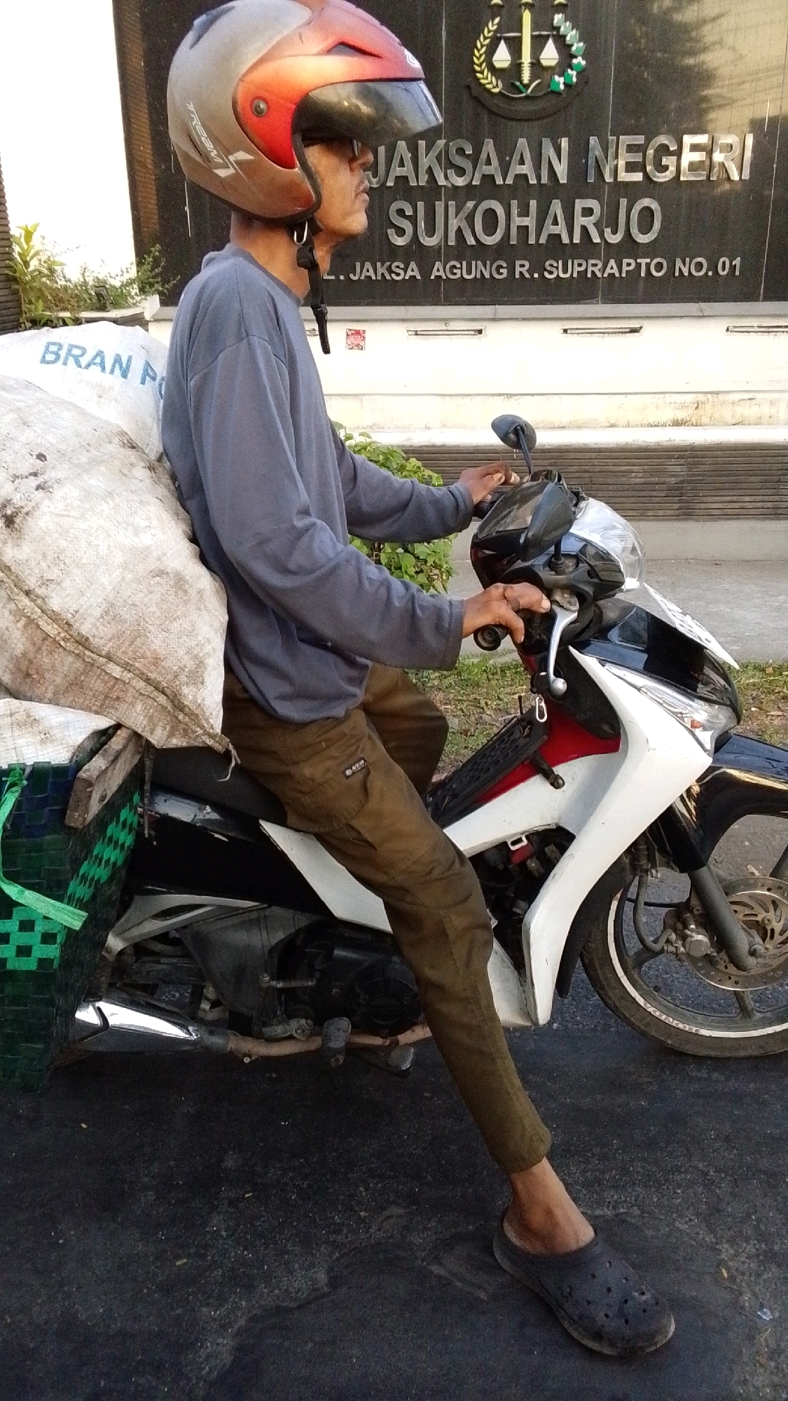 A man wearing a grey long-sleeved shirt, olive green pants, and a dark-colored clog is sitting on a motorcycle. He has a red and silver helmet with a dark visor on his head. The motorcycle is black, white, and red. It is carrying a large, rough, light-colored bag strapped to the back and a green woven basket. The man is looking to his right. Behind the motorcycle is a dark-colored building with a sign that reads "JAKSAAN NEGERI SUKOHARJO" and "L. JAKSA AGUNG R. SUPRAPTO NO. 01." The building has a seal above the text. The ground is asphalt with some debris. The location is identified as Pabelan, Indonesia.