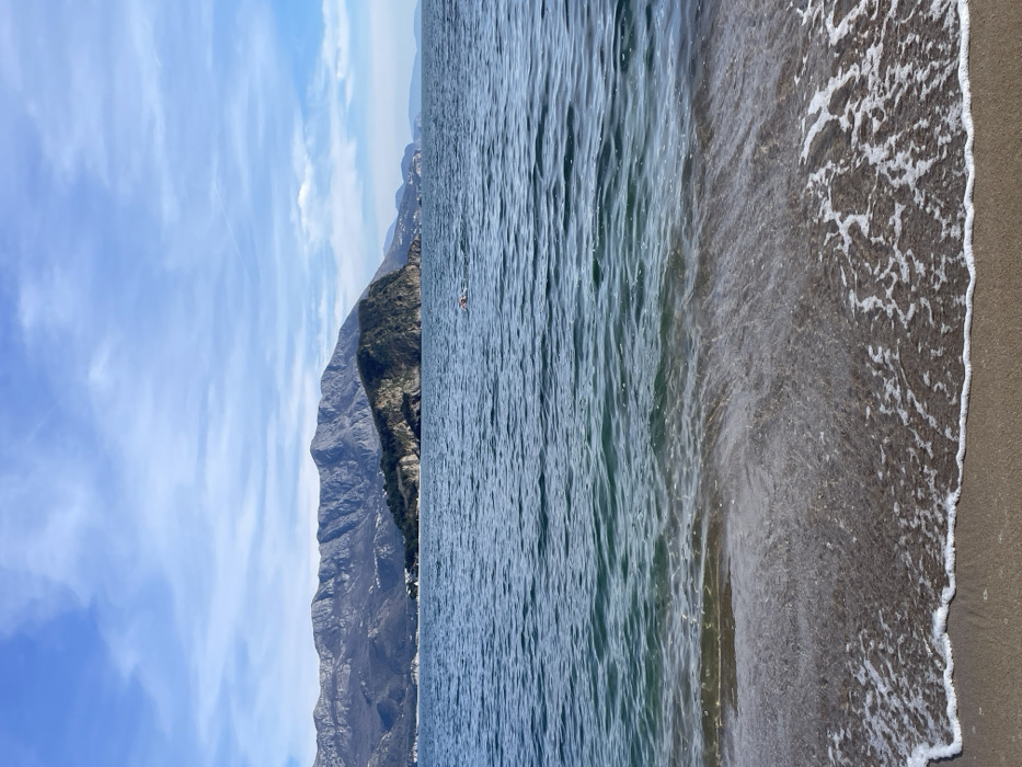 The scene depicts a coastal landscape in Budva, Montenegro. In the foreground, a light brown sandy beach is visible, with shallow, transparent water washing onto the shore, forming white foam. The midground is occupied by a calm expanse of blue-green sea with visible ripples. A single individual is present in the water, positioned centrally, with their head and shoulders partially exposed above the surface, wearing a red cap while engaged in swimming. The background features the prominent, rocky, tree-covered Sveti Nikola Island. Behind the island, a range of grey mountains with visible snow patches extends across the horizon. The sky is light blue with scattered, thin white clouds.