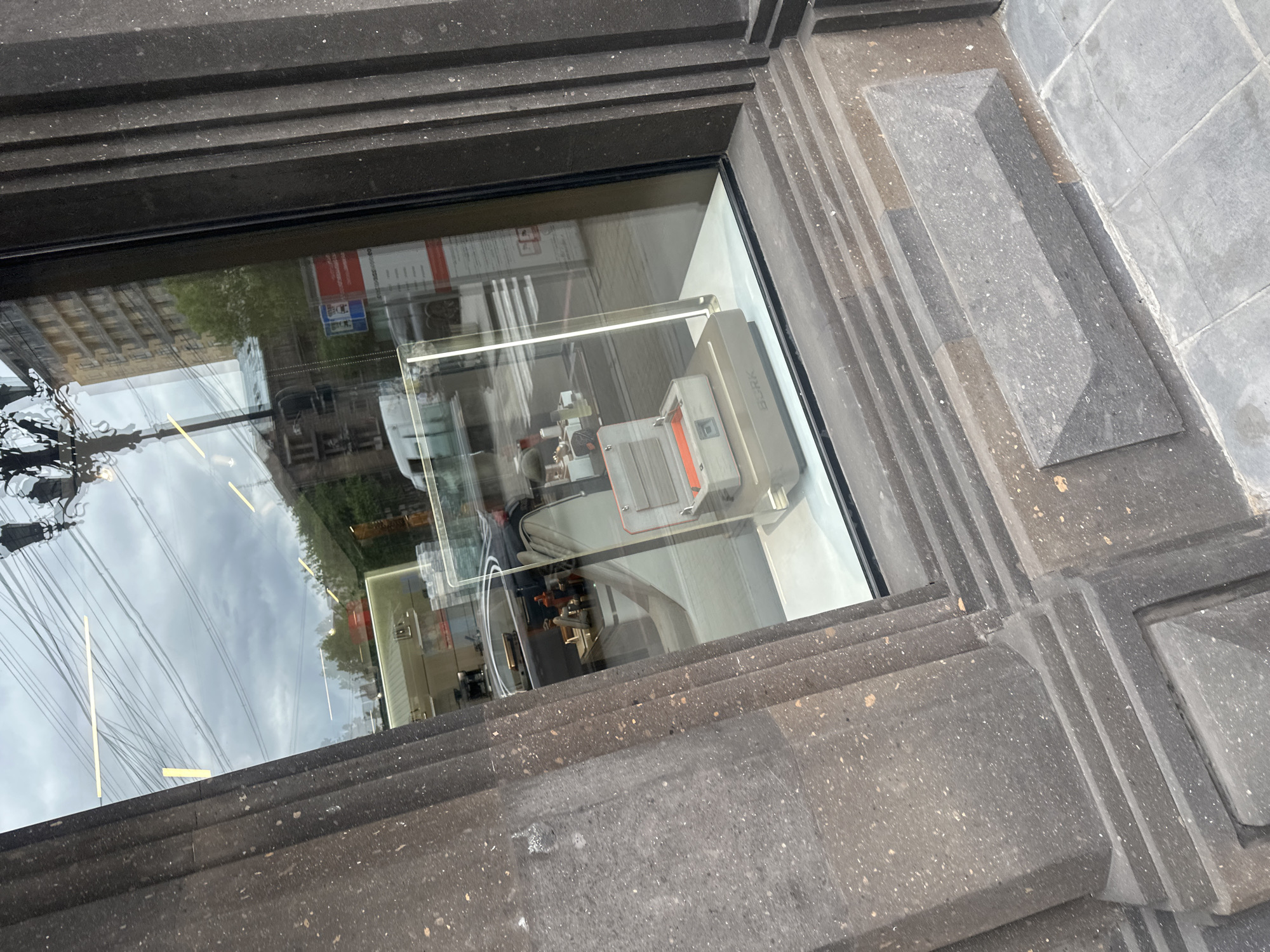The image shows the exterior of a building in Yerevan, Armenia, framed by a window recess constructed of dark, textured stone. The window reflects the exterior environment, including power lines and the facades of other buildings, partially obscured by reflections. Inside the window, a display features a "BORK" branded electrical appliance. The architecture surrounding the window is characterized by geometric, tiered stone elements.