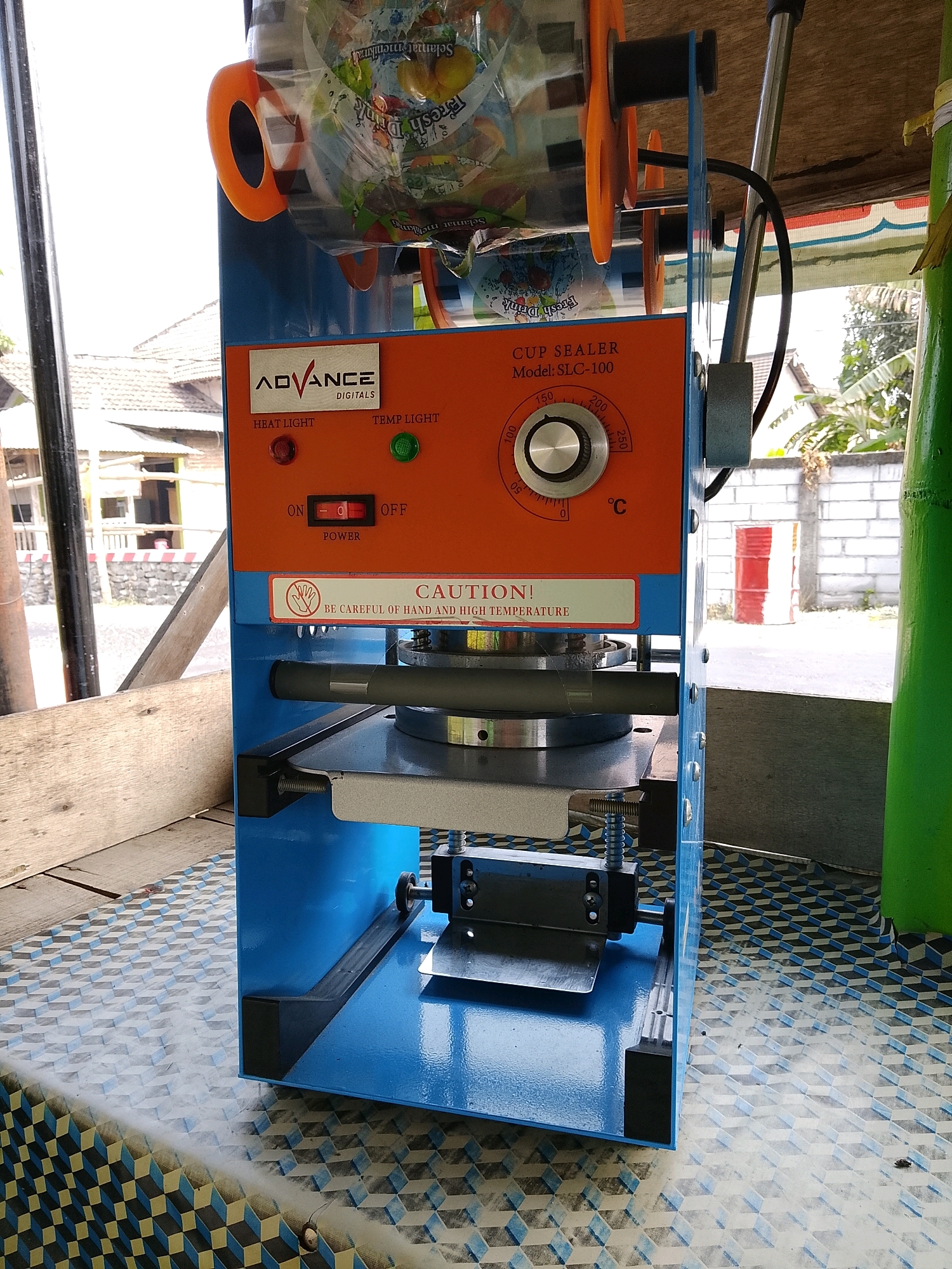 An outdoor scene in Banaran, Indonesia, shows a blue and orange cup sealing machine on a patterned table. The machine has a roll of plastic film with text that appears to be in Indonesian, and a dial with temperature settings. A red heat light and a green temp light are visible on the front of the machine, along with a power switch. A caution label warns about high temperatures. In the background, a building with a tiled roof and a brick wall are present, along with some greenery and a tall green cylindrical object. The lighting suggests it is daytime.