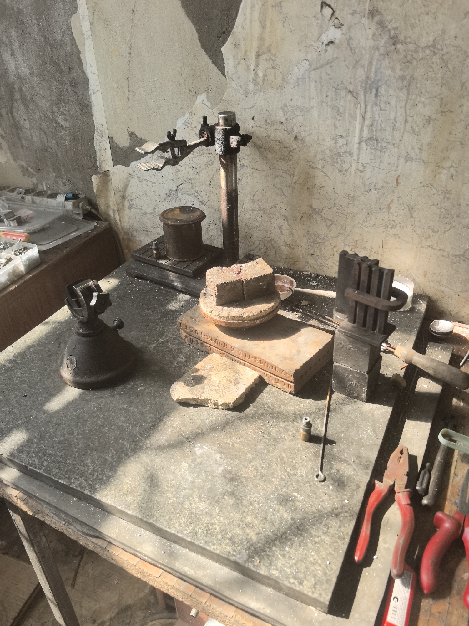 This is a close-up, eye-level, outdoor, still shot of a workbench in what appears to be a jewelry making or metalworking studio. The workbench is cluttered with various tools and materials. The background is a rough, textured wall with peeling paint and plaster. The lighting suggests it is daytime, with strong shadows cast across the scene.
On the workbench, in the foreground, are a pair of red-handled pliers, a green tool, and another red-handled tool. Further back, there is a metal vise, a brick mold, and a soldering brick. There are also small metal objects, possibly findings or components, scattered on the surface. A metal stand with adjustable clamps is positioned behind the soldering brick. A cylindrical metal container sits on a base to the left of the stand.
The overall impression is one of a functional, but somewhat untidy, workspace. The absence of people suggests the area is currently not in use. The scene is consistent with a workshop that might be found in an industrial or artisan district of a city like Amursk, Russia.
