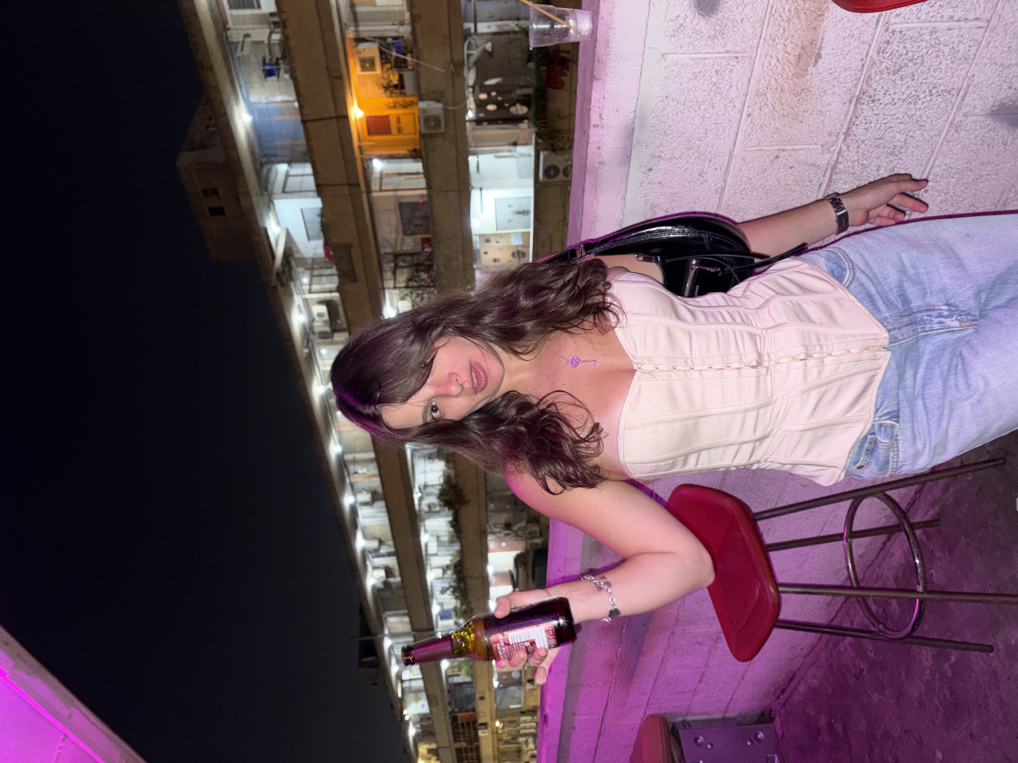 The image depicts a young woman standing at night on what appears to be a rooftop or elevated outdoor bar setting in Tel-Aviv, Israel. She is positioned slightly to the right of the frame's center, holding a brown glass beer bottle in her raised left hand. The woman has long, dark brown hair and is wearing a light beige, strapless corset top, light blue denim jeans, a silver watch on her left wrist, and a silver necklace with a key-shaped pendant. A black shoulder bag is visible on her right side. Her expression involves slightly open lips and a direct gaze towards the viewer.
To her left, a red bar stool with a silver frame is partially visible. The foreground features a low, white, textured concrete wall or bar counter, illuminated by a strong pink/purple neon light source from above, casting a magenta hue across the wall and the floor. On the far right of this wall, a clear plastic cup with a straw is placed.
In the background, under a dark night sky, multi-story residential buildings are visible, illuminated by various interior lights (some warm yellow, some white) from their windows and balconies. Numerous air conditioning units are mounted on the exterior walls of these buildings, which exhibit a somewhat aged, functional architectural style consistent with urban development in Tel-Aviv. The overall atmosphere suggests a lively evening setting in an urban environment.