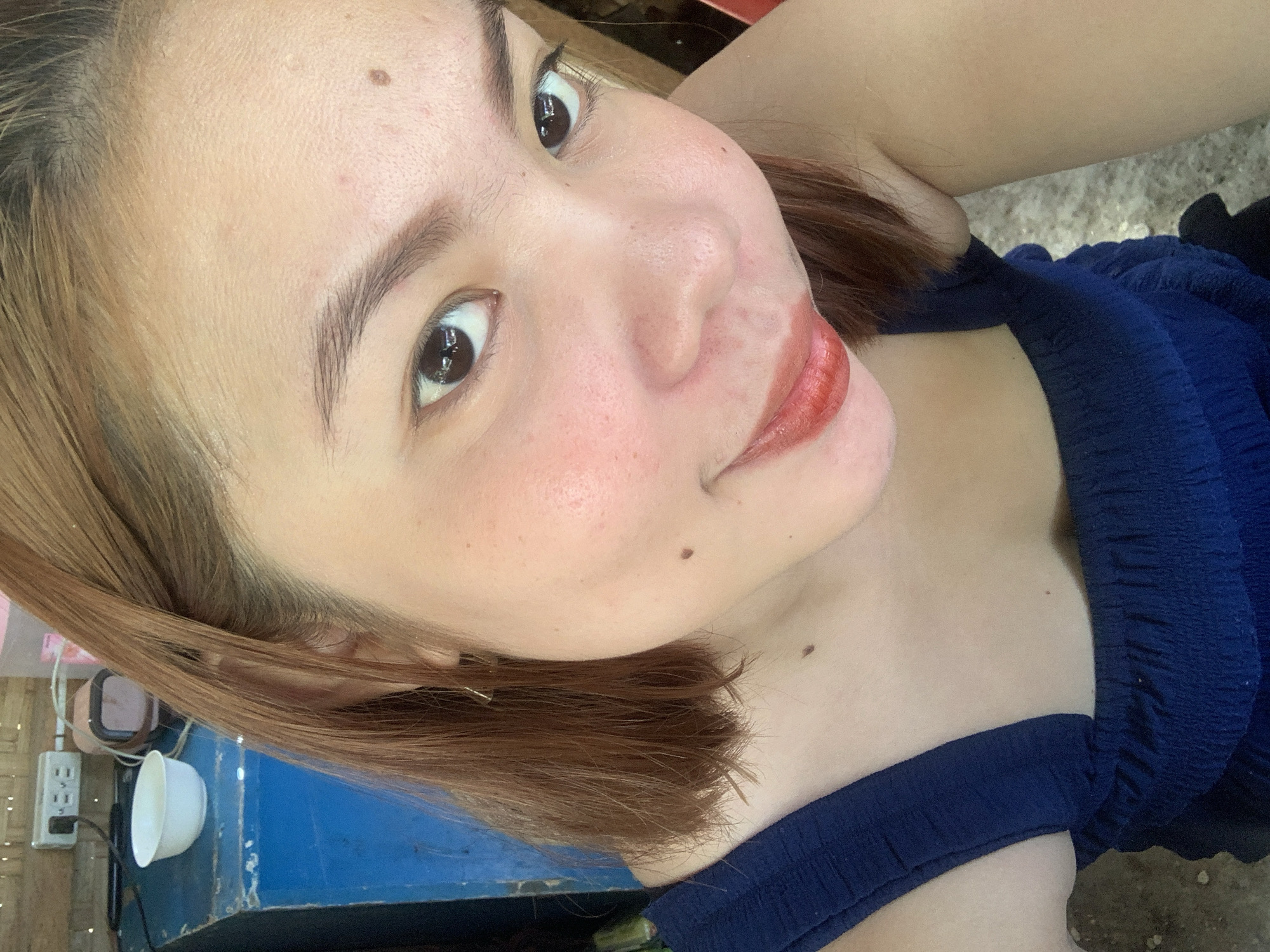 The image shows a close-up selfie of a young woman with light brown hair and fair skin, wearing a dark blue top. She is looking directly at the camera with a slight smile. Her makeup includes red lipstick and blush on her cheeks. There are a few moles visible on her face and chest. In the background, slightly out of focus, is a blue wooden surface, a white bowl, an electrical outlet, and some other objects that are not clearly identifiable. The image appears to have been taken indoors, possibly in a residential setting. There is no specific indication in the image to place it in Kiblawan, Philippines.