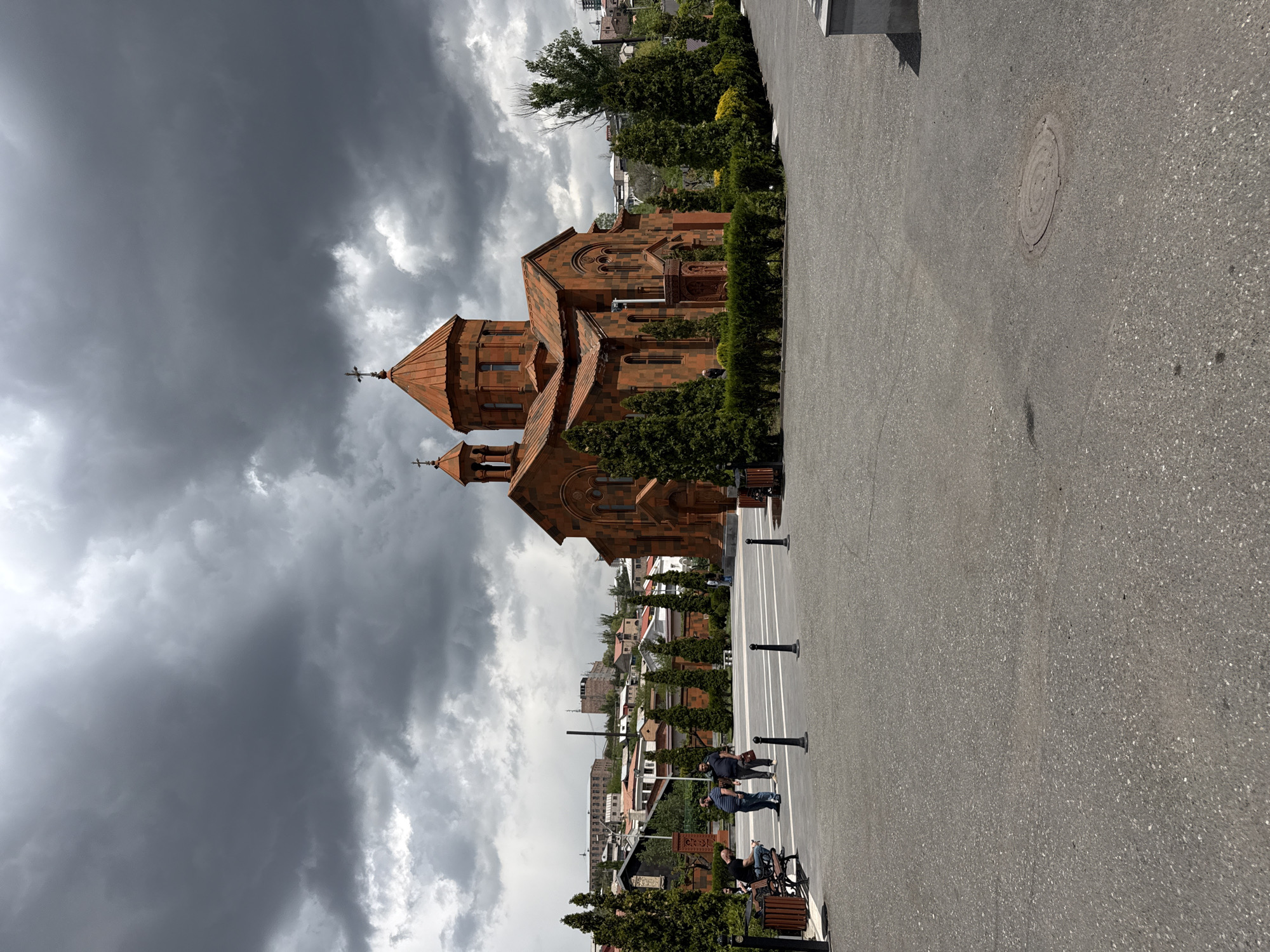 The image shows a building in Yerevan, Armenia. The building is a red-brick structure with a cross on top, possibly a church or cathedral. The sky is overcast with dark, stormy clouds. People are present, walking and sitting on benches. The surrounding area appears to be a paved square. Trees and shrubs border the building. The rest of the city is visible in the background. The image is vertical.