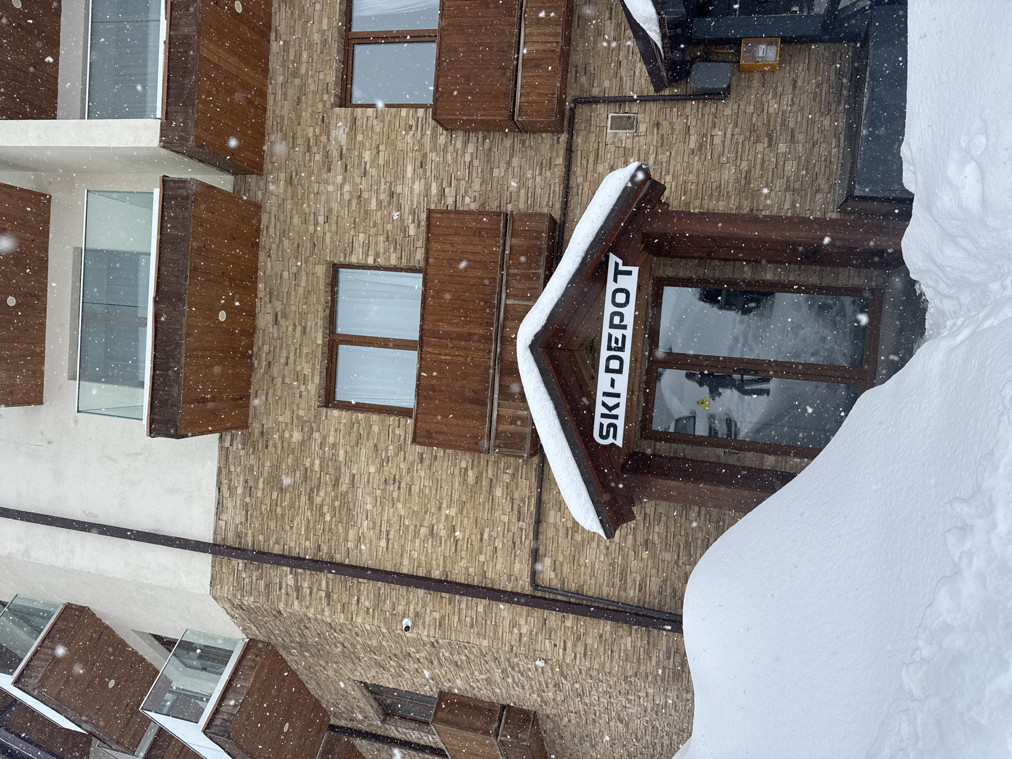The image shows the exterior of a building in what appears to be a snowy environment. The sign reads "SKI-DEPOT." The building has a stone-like facade with windows and brown wooden shutters or accents. There is a significant accumulation of snow in the foreground, obscuring part of the building. The scene suggests a winter location, possibly a ski resort area.