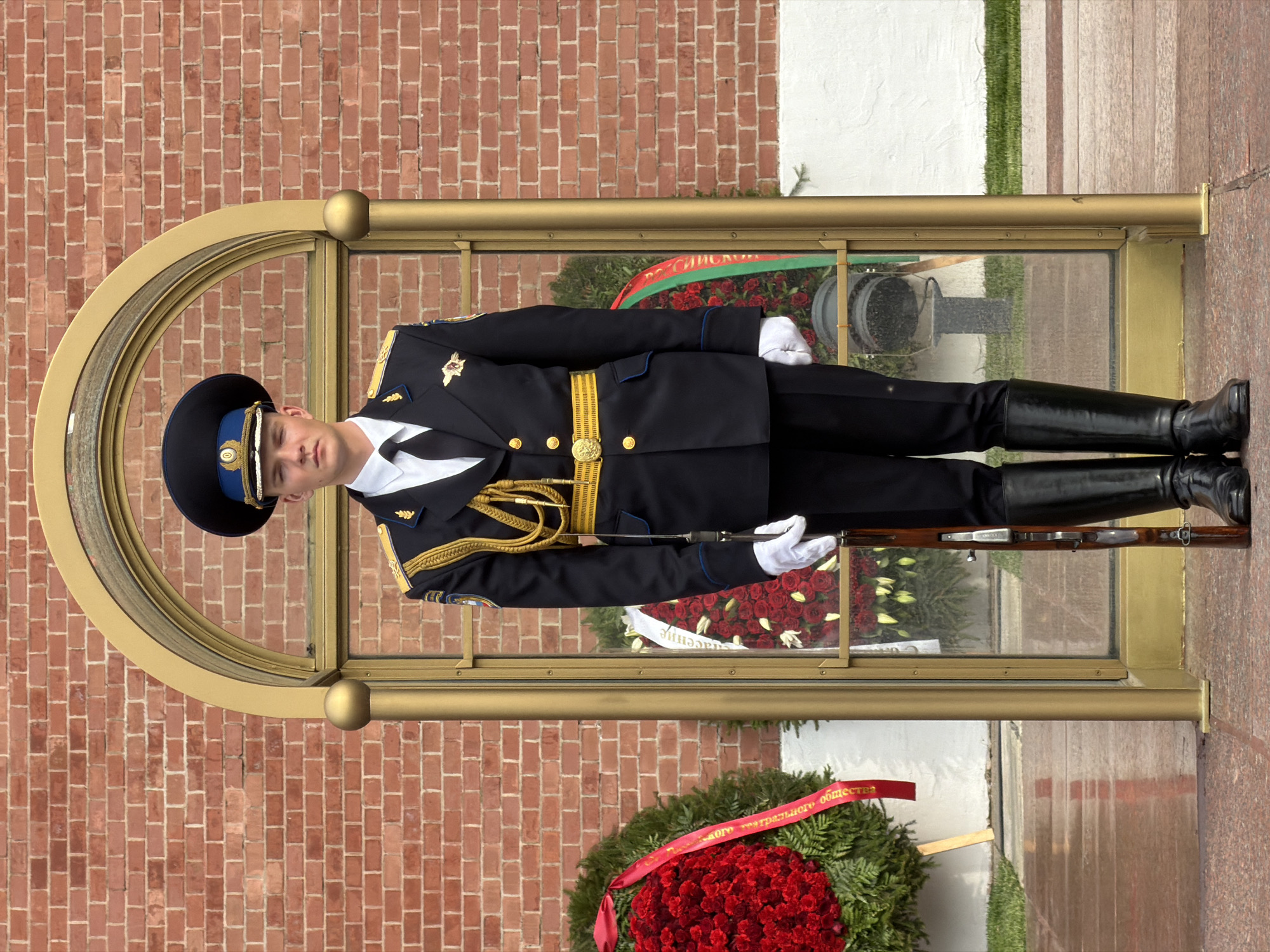 A soldier in ceremonial dress stands guard in front of the Tomb of the Unknown Soldier in Moscow, Russia. The soldier wears a navy blue uniform with gold accents, a black hat, and white gloves. He holds a rifle. The tomb is encased in a glass and gold frame. In the background are wreaths of red roses and greenery, and a brick wall.