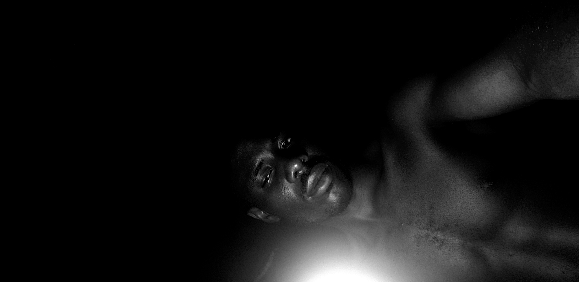 A black and white, low-angle, close-up photograph shows a Black man's head and upper chest against a black background. The man's head is tilted upwards and to his right, with his eyes looking directly at the camera. His skin is glistening with sweat, and he has a mustache and a faint beard. A bright light source is visible to the left of the frame, illuminating his face and chest. The man is shirtless, revealing his chest and shoulder. The image appears to have been taken indoors, possibly in a dimly lit room. No other people or objects are visible in the frame. The scene is set in Narayi, Nigeria.