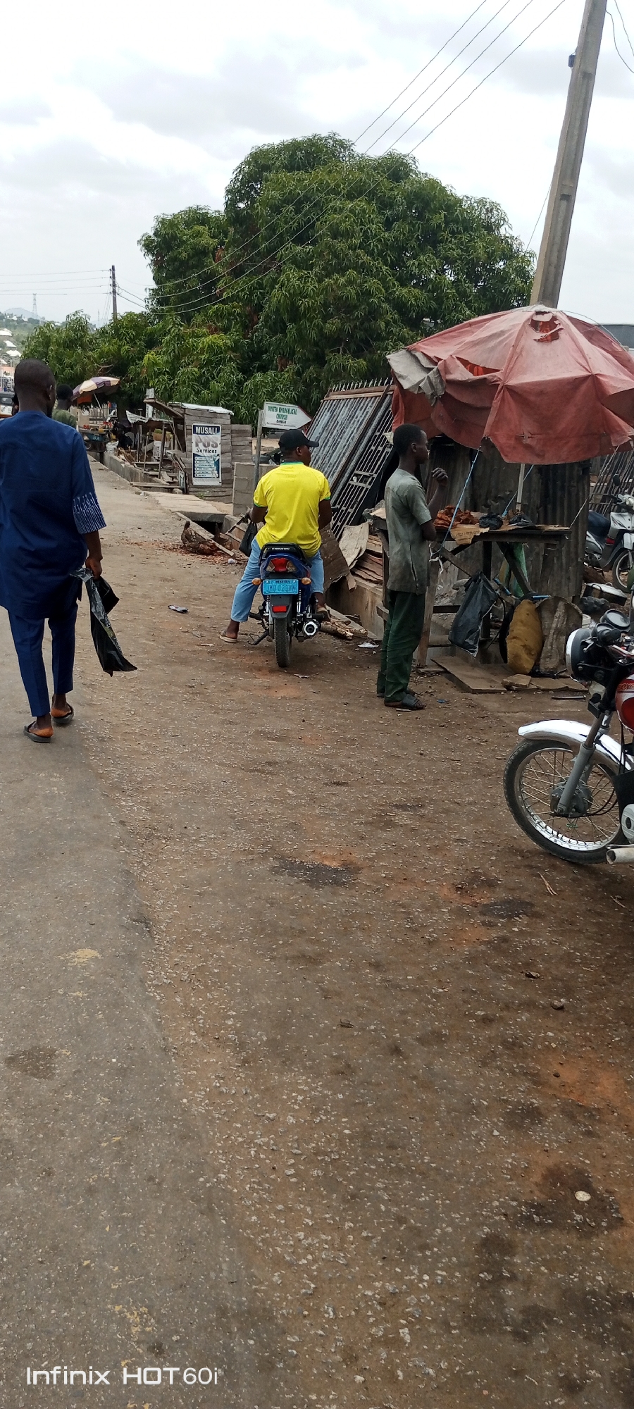The image shows a street scene in what appears to be Zango-ndaji, Nigeria. A man in a blue outfit is walking away from the camera on the left. A man wearing a yellow shirt and blue jeans is sitting on a motorcycle. Another man in green clothes stands near a stall with a torn red umbrella. In the background, there are more stalls and shops, including one with a sign saying "Musali P.O.S. Services." A tree dominates the center background. A partially visible motorcycle sits on the right. The road surface is weathered. An inscription "Infinix HOT 60i" appears in the bottom left corner.