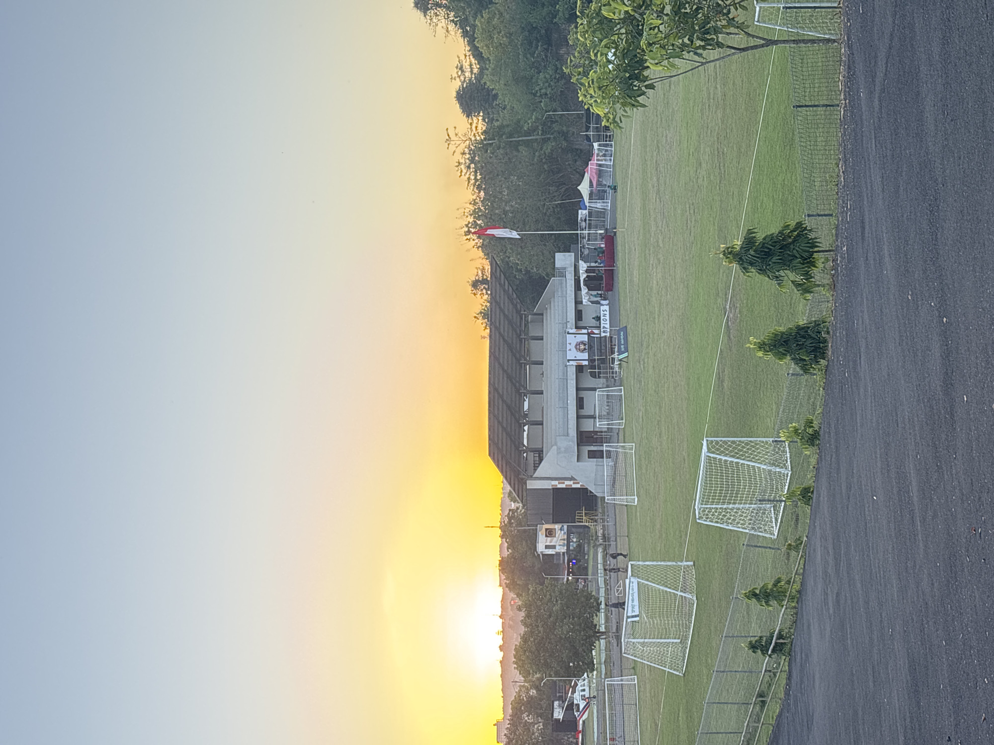The image shows a soccer field in Kuta Selatan, Indonesia at sunset. The sky is transitioning from blue to orange. A grandstand with a roof stands next to the field, with the Indonesian flag flying atop it. Several white soccer goals are placed around the field. Trees line the edges of the field and a black asphalt path runs along one side. The sun is low on the horizon, creating a bright glow.