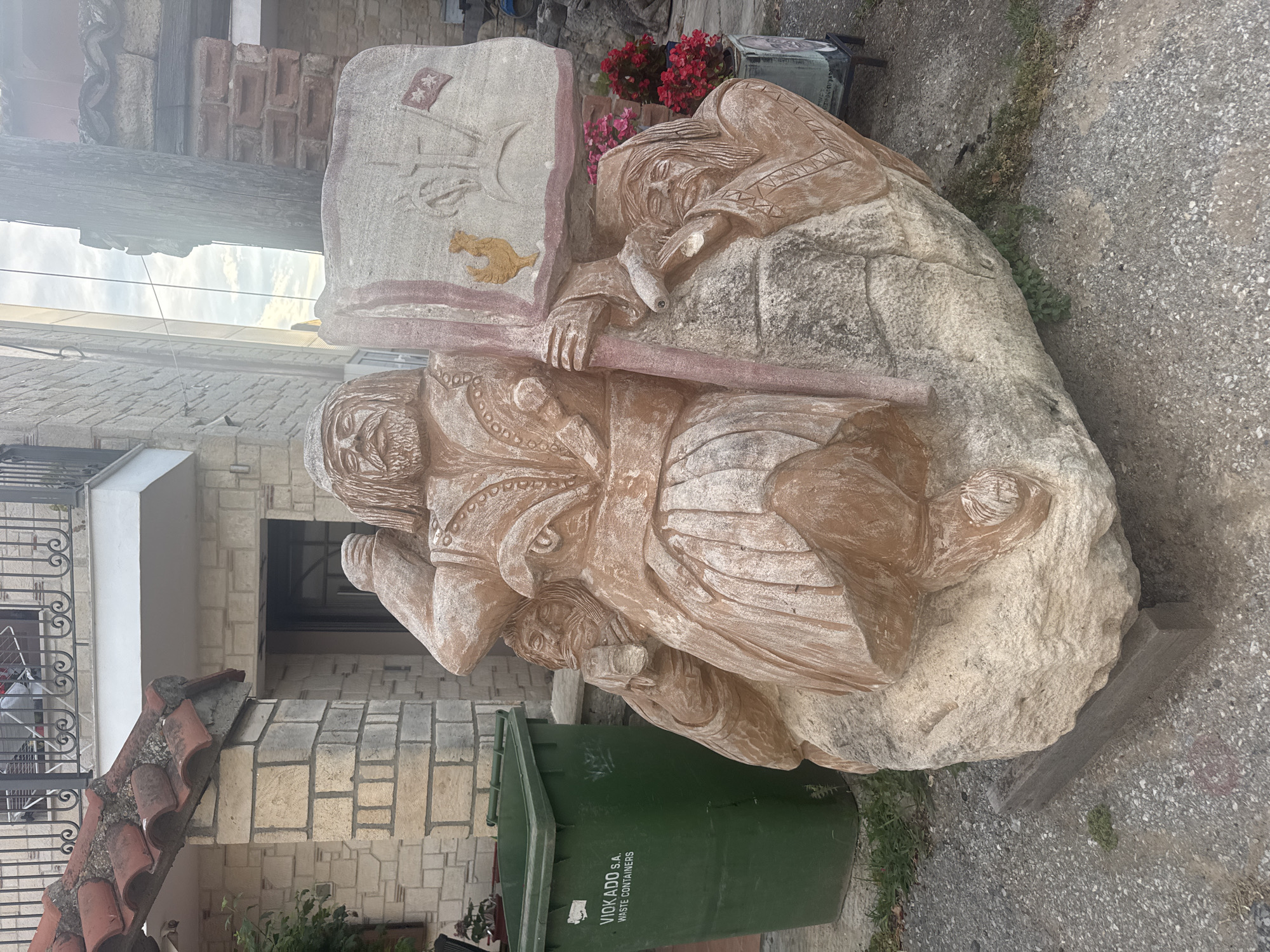 The image shows a stone sculpture depicting a warrior-like figure holding a banner aloft. The sculpture is situated outdoors, in what appears to be a public space or courtyard. The warrior has a long beard and is dressed in ornate clothing. The banner features relief carvings, including a rooster and some symbols. Two smaller, partially obscured faces are carved into the rock at the base of the main figure. To the left of the sculpture is a large green waste container with "VIOKADO S.A. WASTE CONTAINERS" printed on it. Behind the sculpture, a building with stone and tile construction is visible, along with a metal railing. The ground is covered in gravel. In the background, vegetation and a wooden pole are present. The overall scene suggests a rustic or traditional setting. The location is identified as Kassandra Municipal Unit, Greece.