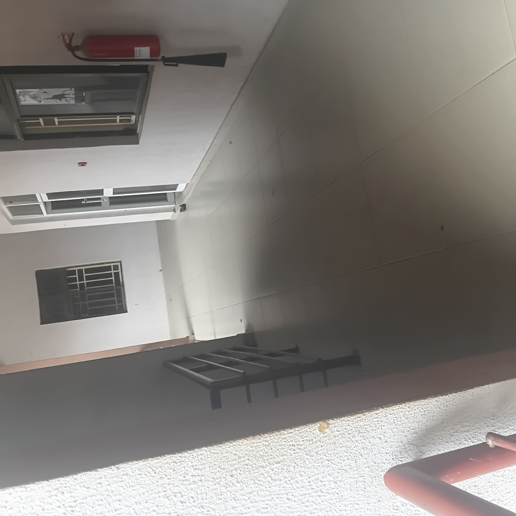 This is a photograph taken indoors, likely in a hallway or common area of a building. The image is oriented sideways, with the camera looking upwards. The scene features a red fire extinguisher mounted on a white ceiling. Below the ceiling, there's a large rectangular opening, possibly a window or a skylight, with visible internal framing. Further down, there's a smaller rectangular window with a metal grate on the outside. The walls appear to be a light color, possibly white or cream, and are tiled in large, rectangular sections. In the foreground, a textured white surface is visible, suggesting a wall or floor finish, and a portion of a red pipe or railing is present. The lighting suggests it might be daytime, but the indoor setting makes it difficult to determine the exact time or weather conditions. There are no people visible in the image. No legible text is visible.