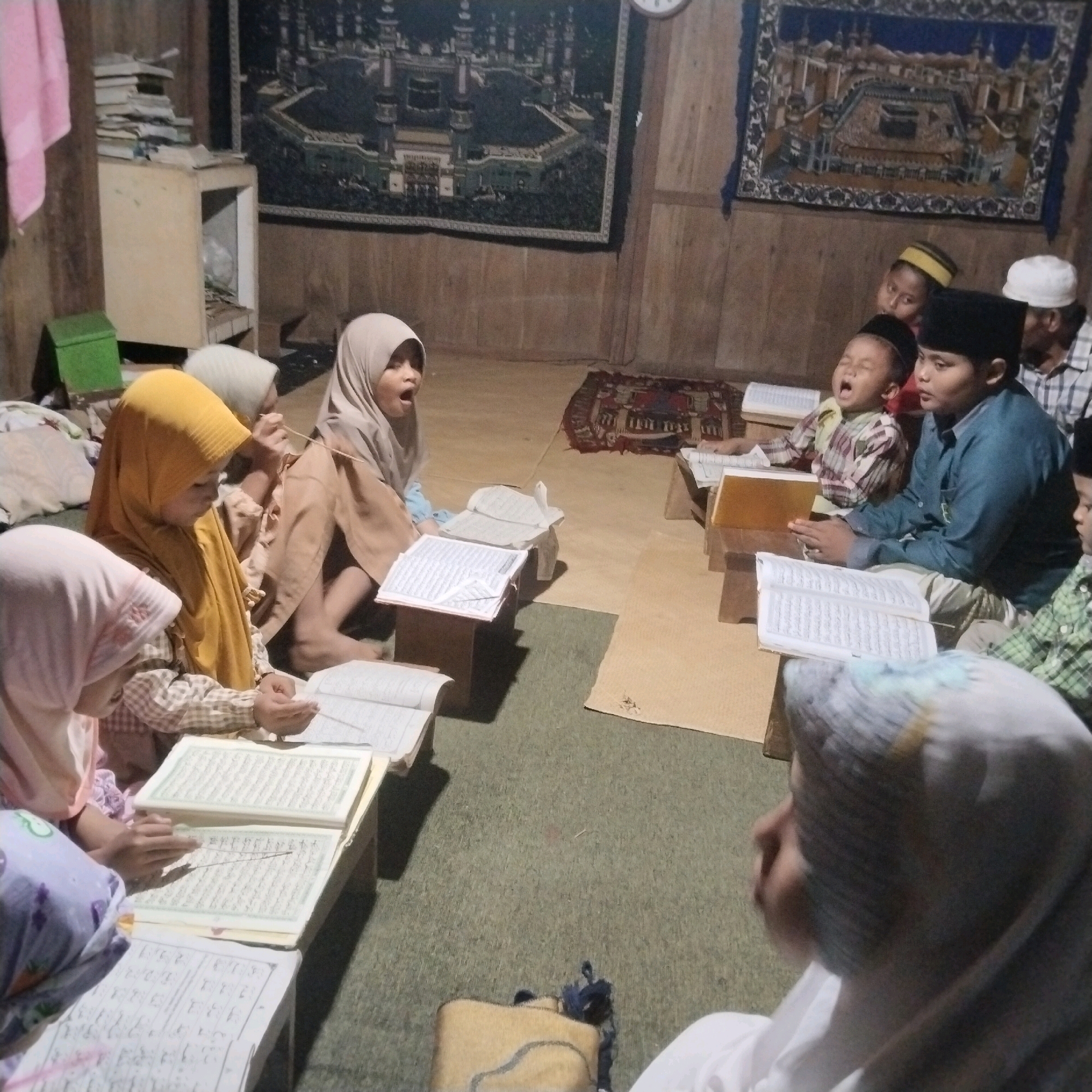 In Banyubunih, Indonesia, a group of children are gathered in what appears to be a communal learning space. The room has a rustic feel, with wooden walls and a carpeted floor. A tapestry depicting a Middle Eastern city, possibly Mecca, hangs on the wall, suggesting a religious context.
Several children are seated on the floor, engrossed in studying religious texts. One girl, wearing a yellow hijab, is attentively reading a book, while another child in a grey hijab is looking up and appearing to speak or sing. In the background, other children are also engaged in their studies, some wearing traditional caps. An older man in a checkered shirt and a white cap is also present, observing the children.
The environment seems to be a humble dwelling or a modest community center, with simple furniture like wooden stands holding books and what might be a small cabinet. The overall scene conveys a sense of collective learning and spiritual devotion among the young members of the Banyubunih community.