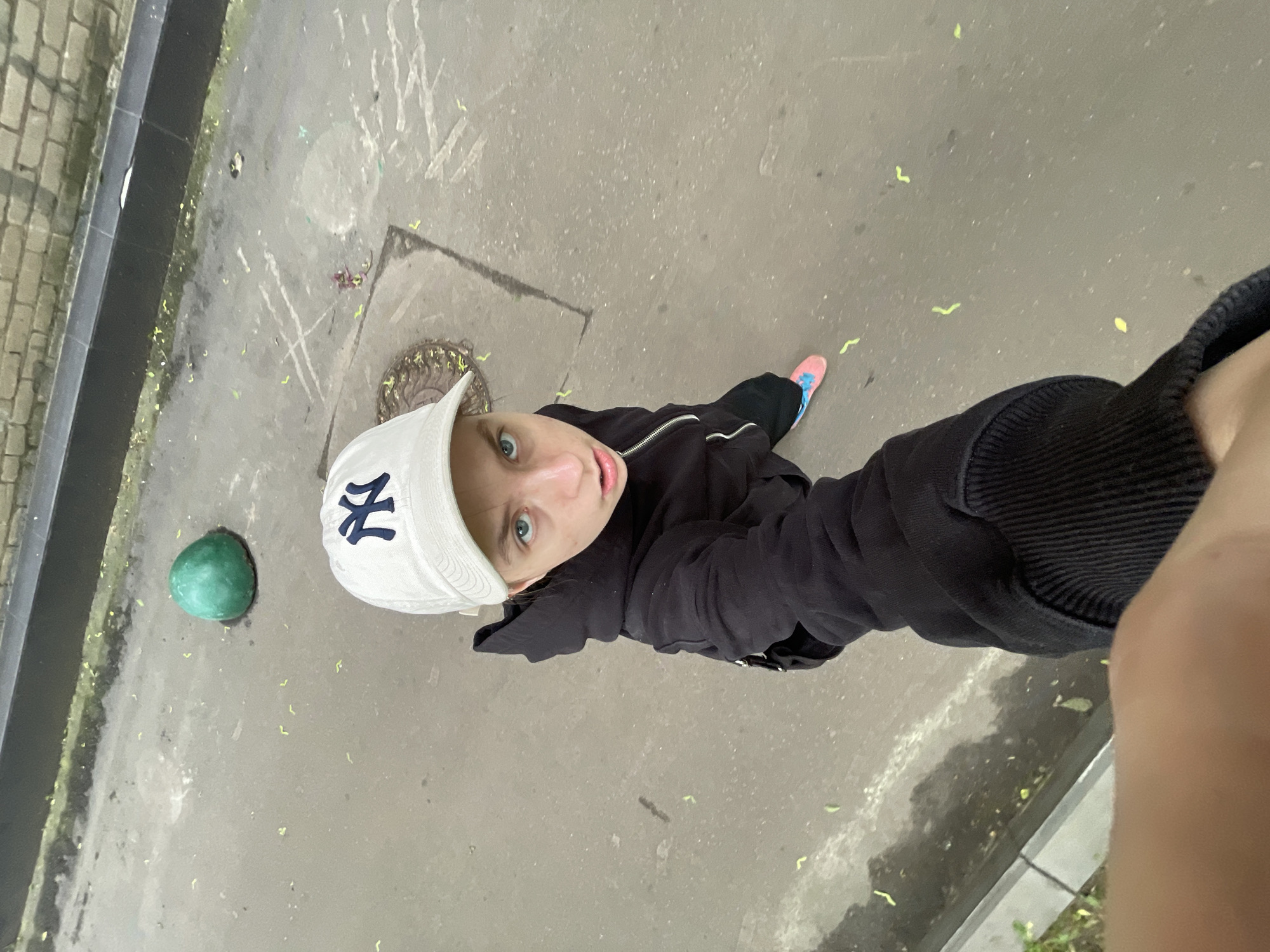 A high-angle view depicts a person wearing a white baseball cap with a stylized navy blue "NY" logo, a black jacket, and dark pants, standing on a concrete surface. One of their feet, clad in a colorful athletic shoe, is visible. The person is looking upwards into the camera. Beside the person is a green spherical object on the concrete. The ground has scattered light debris and is marked with chalk or similar material. A manhole cover is also visible on the surface. The location is likely Moscow, Russia based on the manhole cover and overall urban appearance. A raised ledge or curb is visible on the left and right edges of the frame. A person's arm and hand holding the camera are partially visible on the right.