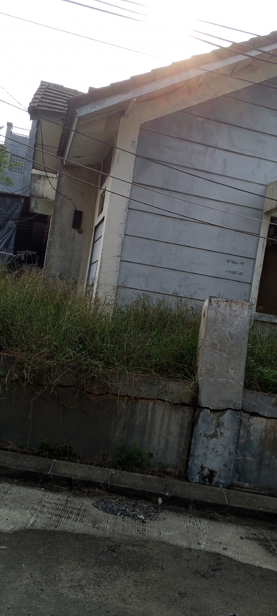 The image shows a weathered, two-story building with light gray siding and a tiled roof, possibly a residence, in Gunung Putri, Indonesia. The building appears to be situated on a slight incline, with overgrown grass and vegetation growing on a retaining wall in front of it. A concrete pillar stands to the right of the grass, showing signs of age and weathering. The ground in front is paved with asphalt and concrete. Electrical wires are visible running across the top of the building and against the sky. The overall impression is one of a slightly neglected or aged property in a residential area.