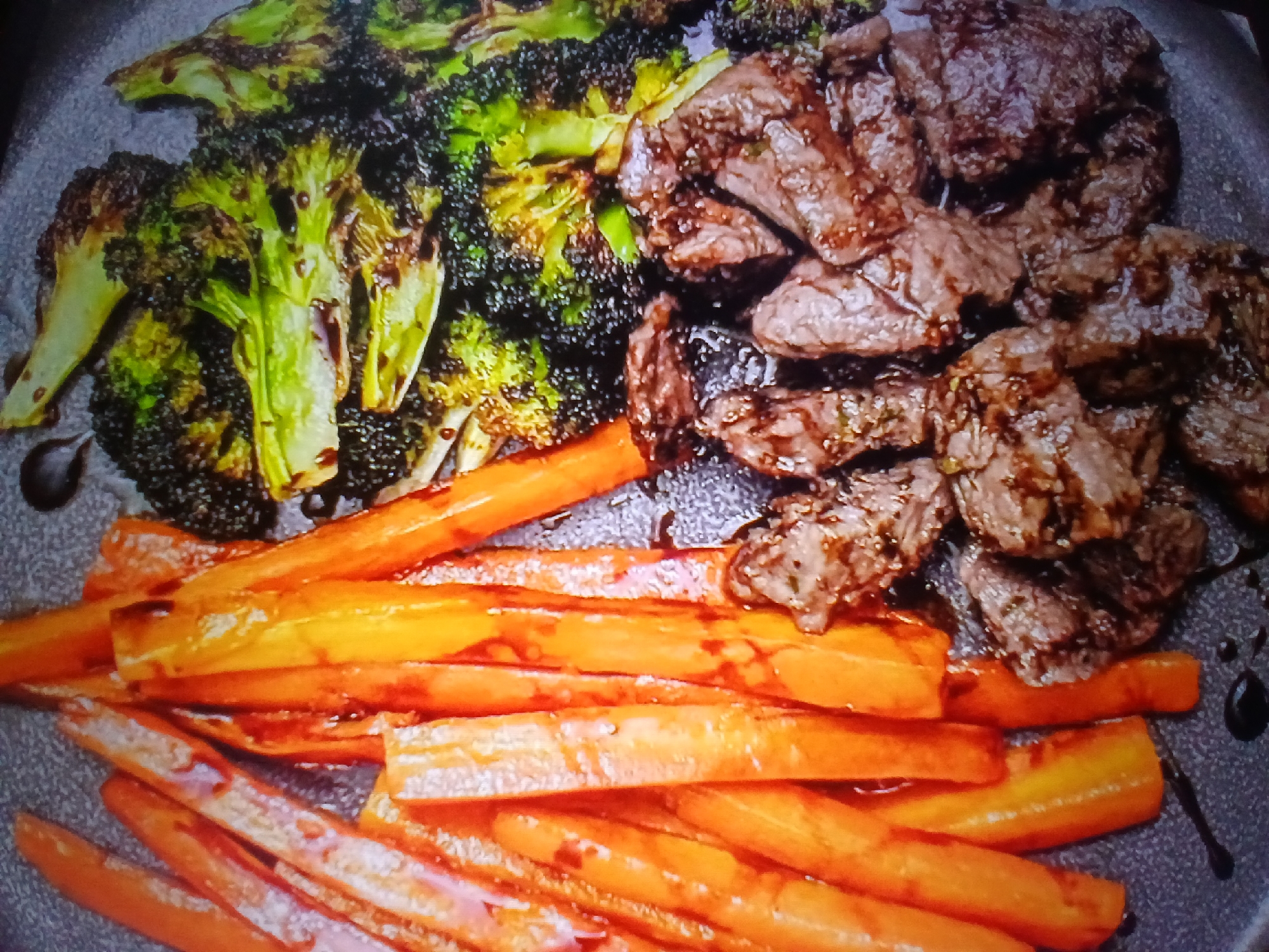This is a close-up, overhead shot of a meal served on a dark plate. The meal consists of three distinct components arranged in sections.
In the upper left quadrant are florets of roasted broccoli, some with charring on the edges, and appearing to be drizzled with a dark sauce, possibly balsamic glaze. The broccoli is a vibrant green with some darker, almost black, burnt tips.
To the right of the broccoli is a pile of sliced, cooked beef. The beef pieces are irregular in shape, and appear to be pan-seared or grilled, with some visible grill marks. They are coated in a dark, glossy sauce.
The bottom portion of the plate is filled with sliced carrots. These appear to be roasted or sautéed, and are a bright orange color, with some lighter, almost white, dusted on them, possibly powdered sugar or salt.
The overall impression is a well-prepared and appetizing meal. There is no indication of the time of day, weather, or specific location beyond the general context of a meal being served. No people are visible in the frame. There is no visible text.