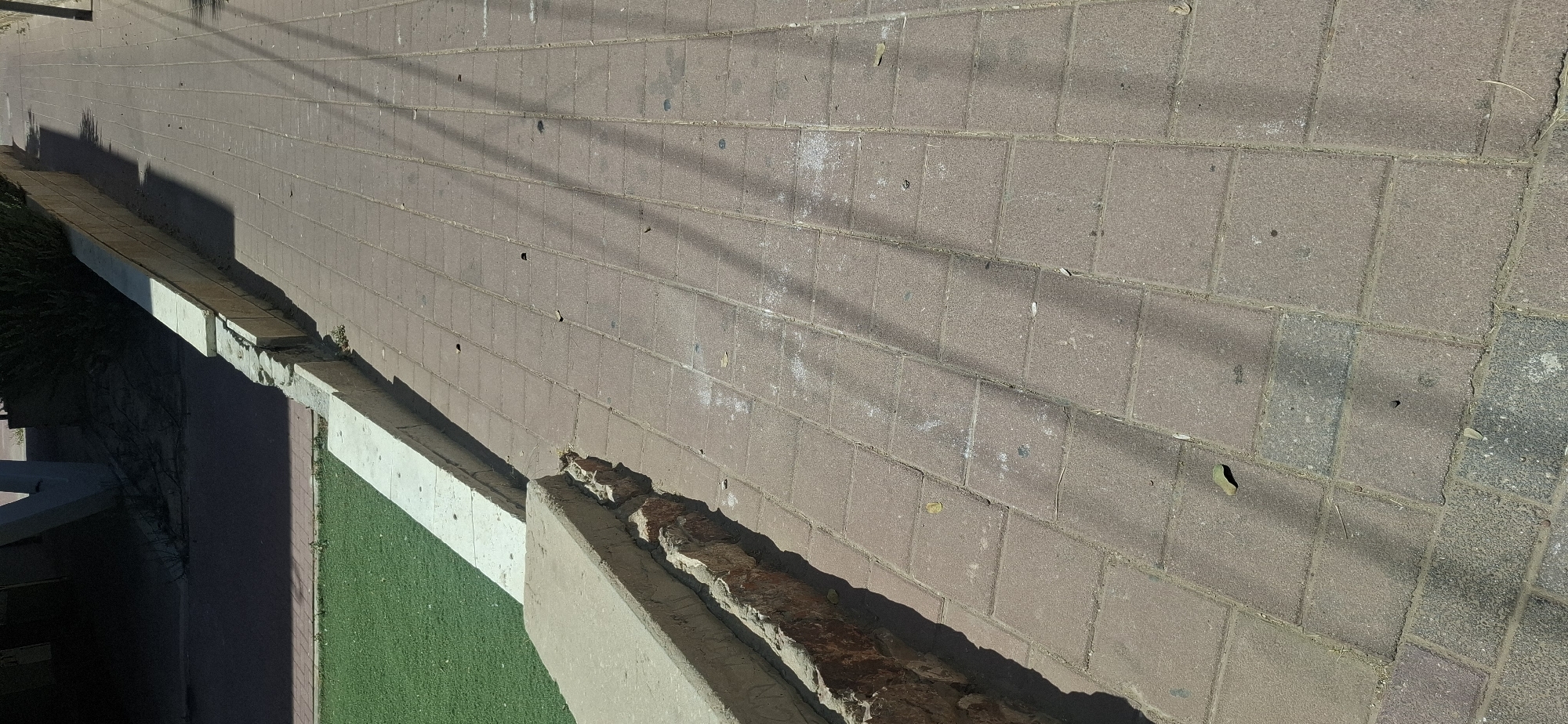 The image shows a sidewalk made of reddish-brown rectangular paving stones. The sidewalk is adjacent to a low wall, which has a section of lighter colored concrete capping. The wall separates the sidewalk from an area of artificial green turf. The wall shows signs of damage and erosion at its base, with exposed reddish-brown rocks and debris. Shadows from an unseen object, likely trees or poles, are cast across the sidewalk. A dark shadow covers the area to the left of the wall, which appears to be a darker paved surface. The overall scene depicts an outdoor urban or public space in Yerocham, Israel, characterized by durable outdoor materials and visible wear.