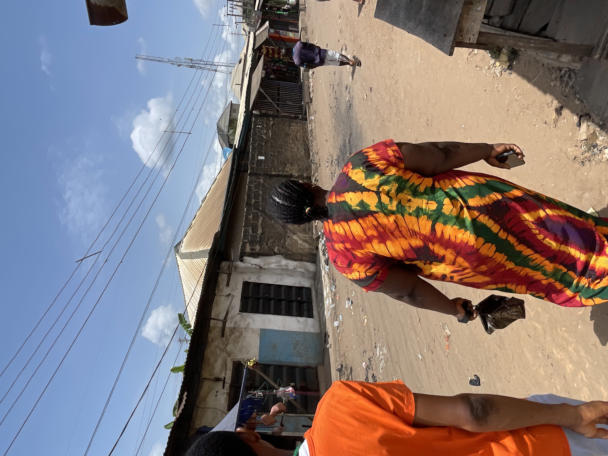 The photograph shows a street scene in Obosi, Nigeria. A woman wearing a colorful, patterned dress is walking along the street, holding a black object in her hand. In the foreground, a person wearing an orange shirt is partially visible. In the background, a man is walking away down the street. Buildings line the street, with corrugated metal roofs and weathered walls. Overhead, power lines are visible against a blue sky with scattered clouds. The street appears unpaved and dusty.