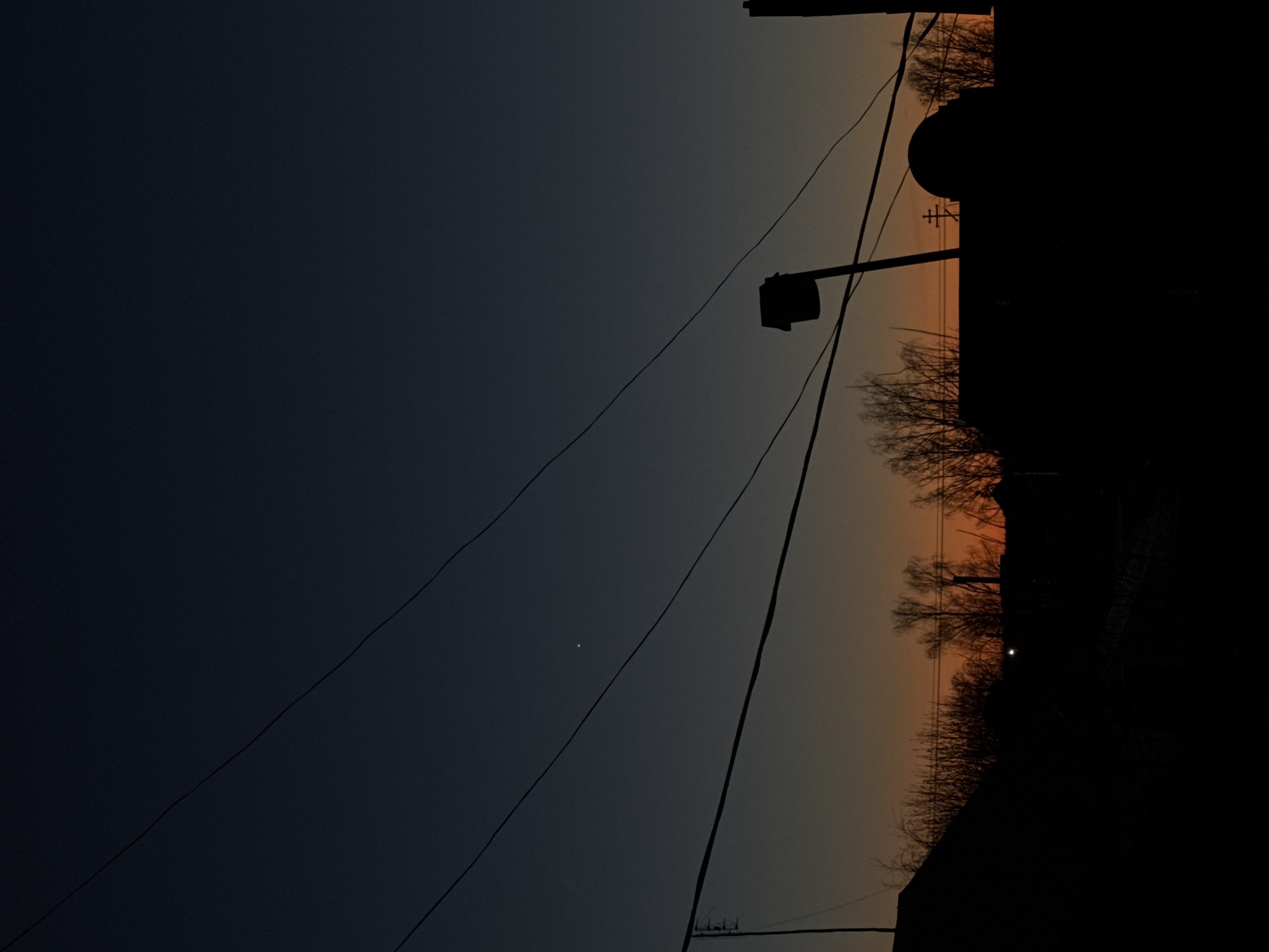 The image shows a dusk scene in Фоки, Russia. Silhouetted against the orange and grey sky is a mix of vegetation, buildings, and utility infrastructure. Trees with bare branches are visible, along with rooftops and what appears to be a water tower or similar structure. Power lines stretch across the sky, intersecting with a street lamp. A small bright spot, possibly a star or planet, is visible in the upper left portion of the sky.