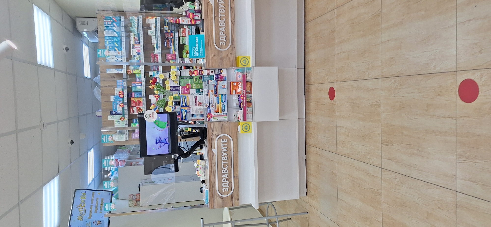 The image depicts the interior of a pharmacy in Nakhodka, Russia. The scene is dominated by a counter with shelves filled with various medicines and health products. A computer monitor is visible on the counter, displaying an animation of two mice. A transparent protective screen is installed on the counter, likely for hygiene purposes. On the counter, the word "ЗДРАВСТВУЙТЕ" (hello in Russian) is prominently displayed on a wooden panel. There are also yellow circular stickers on the floor, indicating social distancing markers. The floor is tiled with a light brown wood-like pattern. The overall environment appears to be clean and organized.