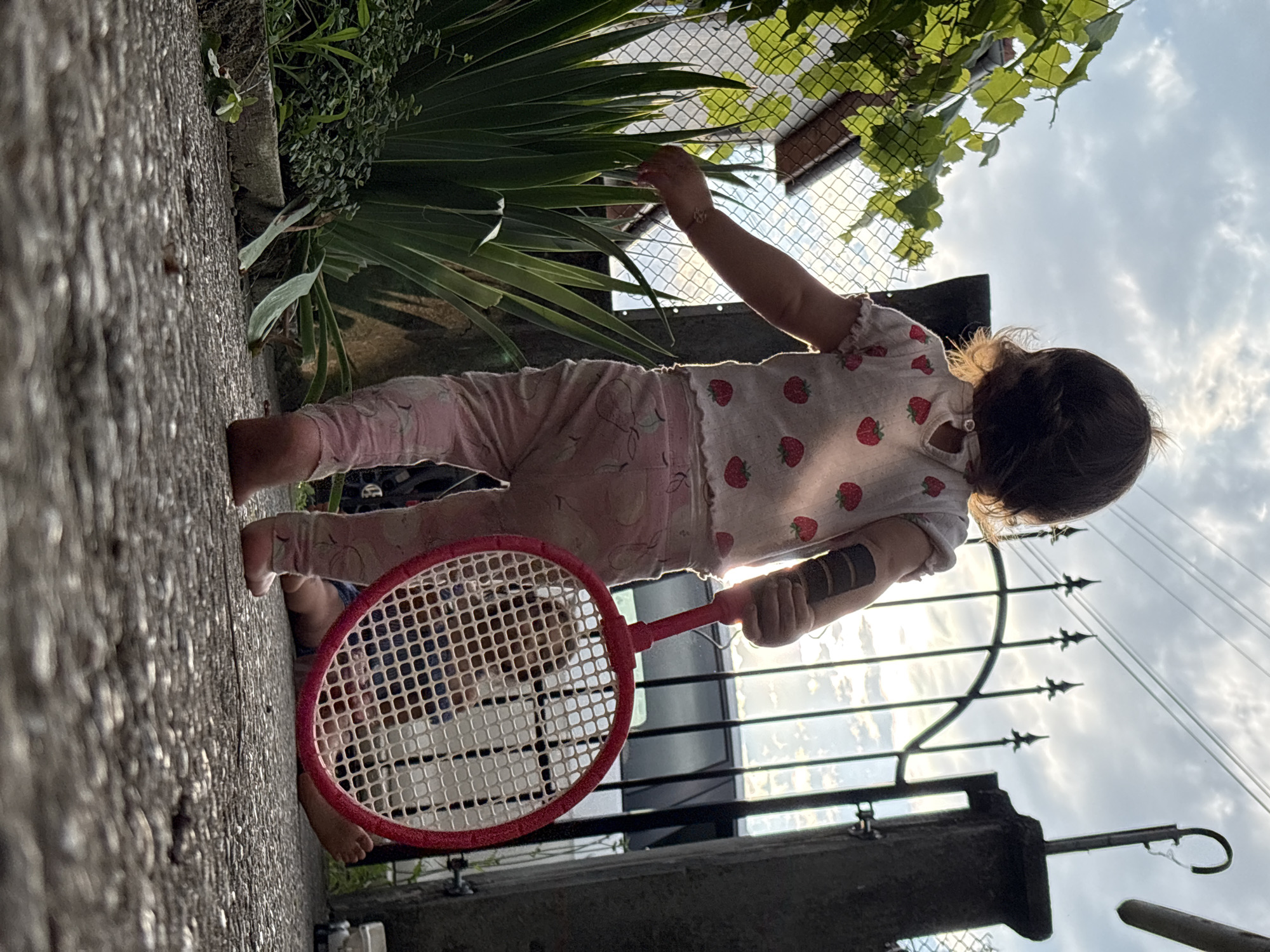 In Oreshak, Bulgaria, a young child stands on a paved walkway, facing away from the camera, holding a child-sized pink badminton racket. The child wears a white t-shirt adorned with strawberries and pink patterned leggings. To the left, lush green plants and a fence with a house in the background are visible. Behind the child, a black metal gate stands open, with a car partially visible through it. Another young child is lying on the ground behind the gate, their legs and torso visible through the racket's netting. The scene is illuminated by natural light, suggesting it is daytime, with clouds present in the sky.