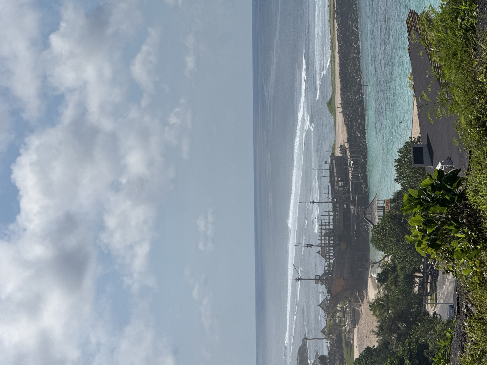 An eye-level, outdoor shot looking out to sea in Ungasan, Indonesia. The sky is overcast. The ocean has rough waves. A structure of wooden poles is present in the distance, near the shore. Beach foliage and trees are in the foreground. A concrete parking lot is visible.