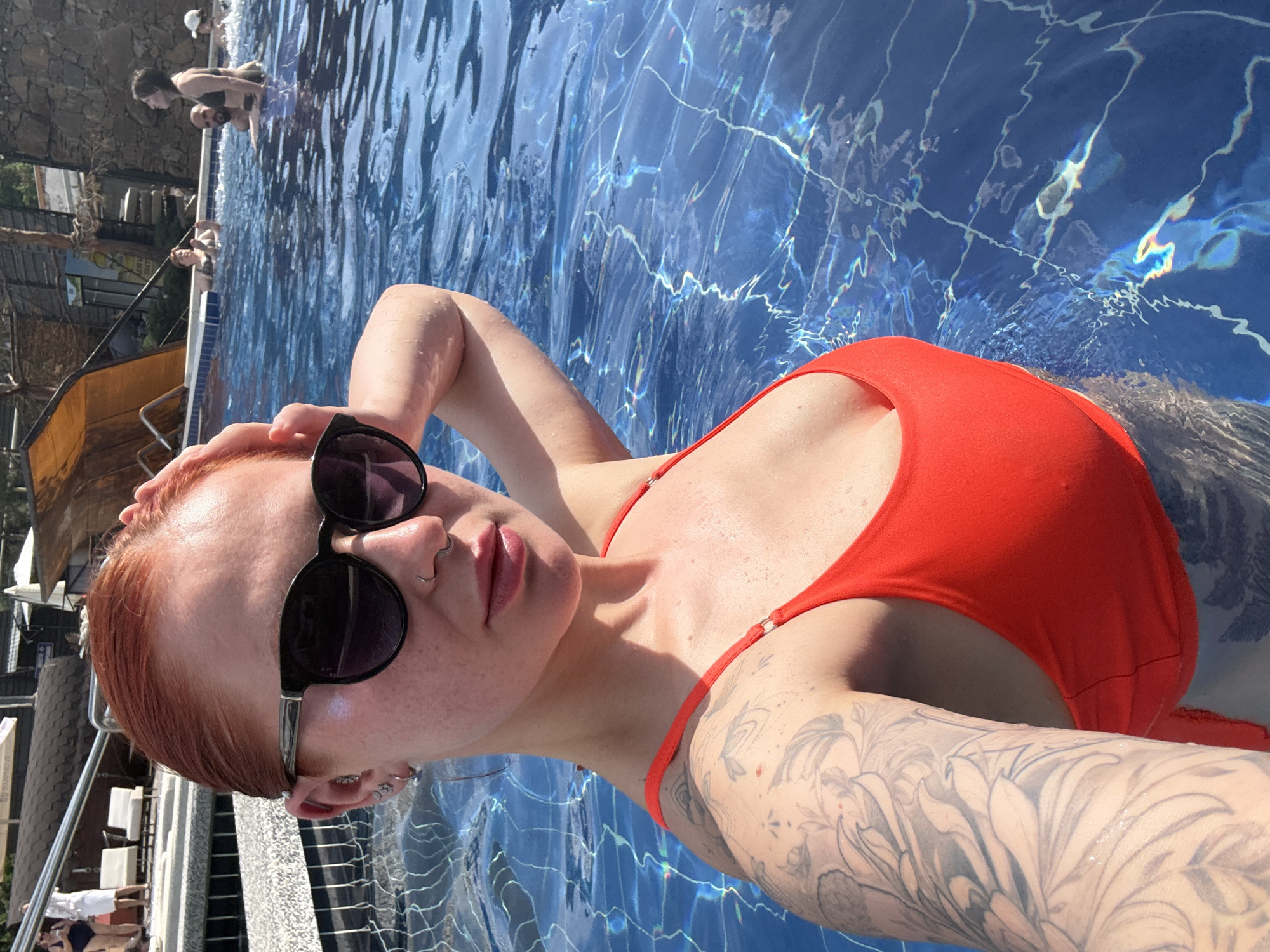 A red-haired woman wearing sunglasses and a red swimsuit is reclining in a blue-tiled swimming pool. The pool water has reflective patterns. The woman has a tattoo on her left arm. In the background, other people are visible near the edge of the pool, and there is a building with white railings and a sloped, wooden structure. The city of Rostov-on-Don, Russia is not directly represented in the image.