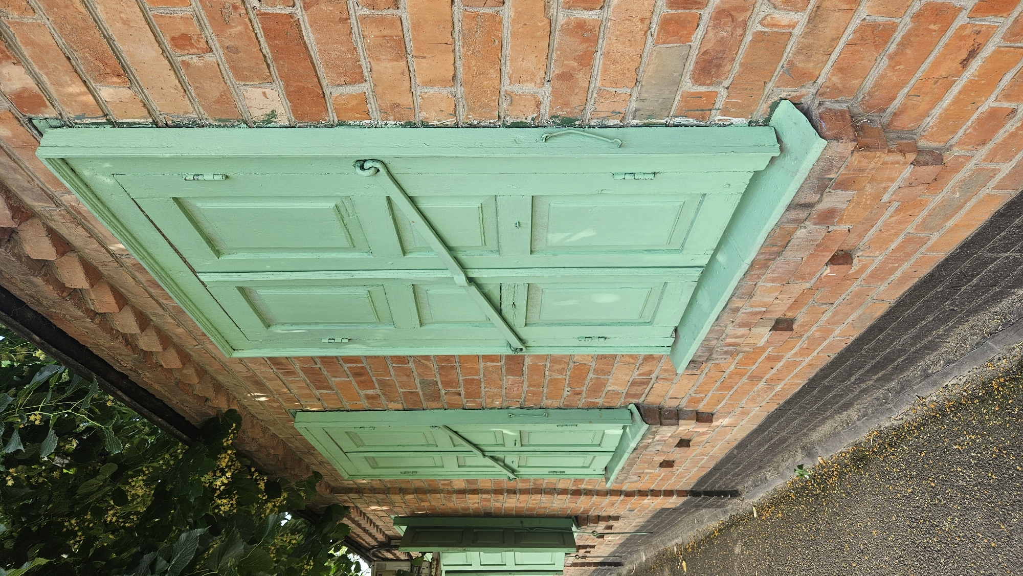 The image shows the exterior of a brick building with two sets of green wooden shutters. The shutters on the right are closed, while the shutters on the left are partially open, revealing a glimpse of the interior. The building has a red brick facade, with a darker colored brick base. A portion of a tree with green leaves and small yellow flowers hangs over the top left of the building. A sidewalk or path made of asphalt runs along the base of the building. The scene appears to be an outdoor view of a residential or commercial property in Krasnodar, Russia.