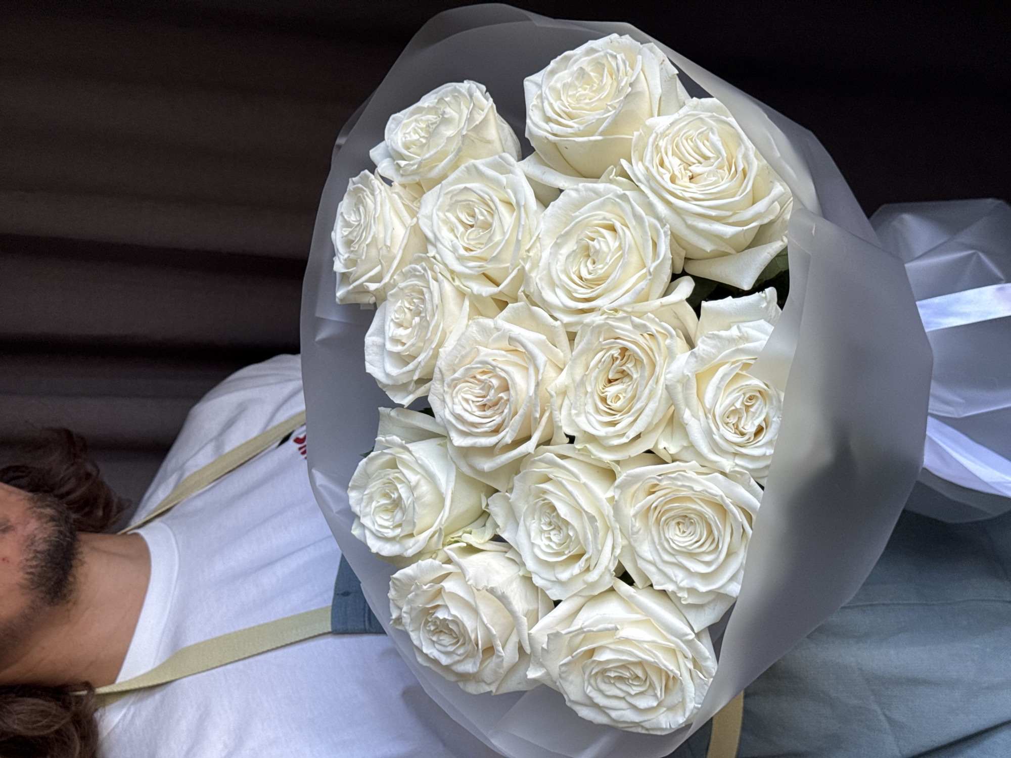 This media file is a close-up shot of a bouquet of white roses. The roses are tightly packed and wrapped in translucent paper. A person is partially visible in the lower left corner, wearing a white t-shirt and appears to be lying down. The person has dark, curly hair and a beard. The background is dark and textured, suggesting an indoor setting or a shaded outdoor area. There is no visible text in the image. The lighting is soft, highlighting the delicate petals of the roses. The overall mood is serene and elegant. The location context is Izhevsk, Russia.