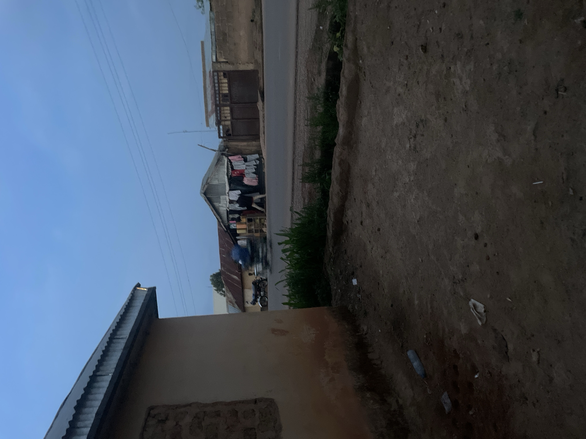 The image shows a street scene in Reifild-Ist, Nigeria. A road runs horizontally across the middle, flanked by buildings on either side. On the left, clothes are hanging outside of a building to dry. A person on a motorcycle is on the road in the direction of the clothing. A building is in the foreground on the left side of the image. In the foreground of the right side is what appears to be a sidewalk that leads to a grass patch beside the road. The sky is visible above the buildings.