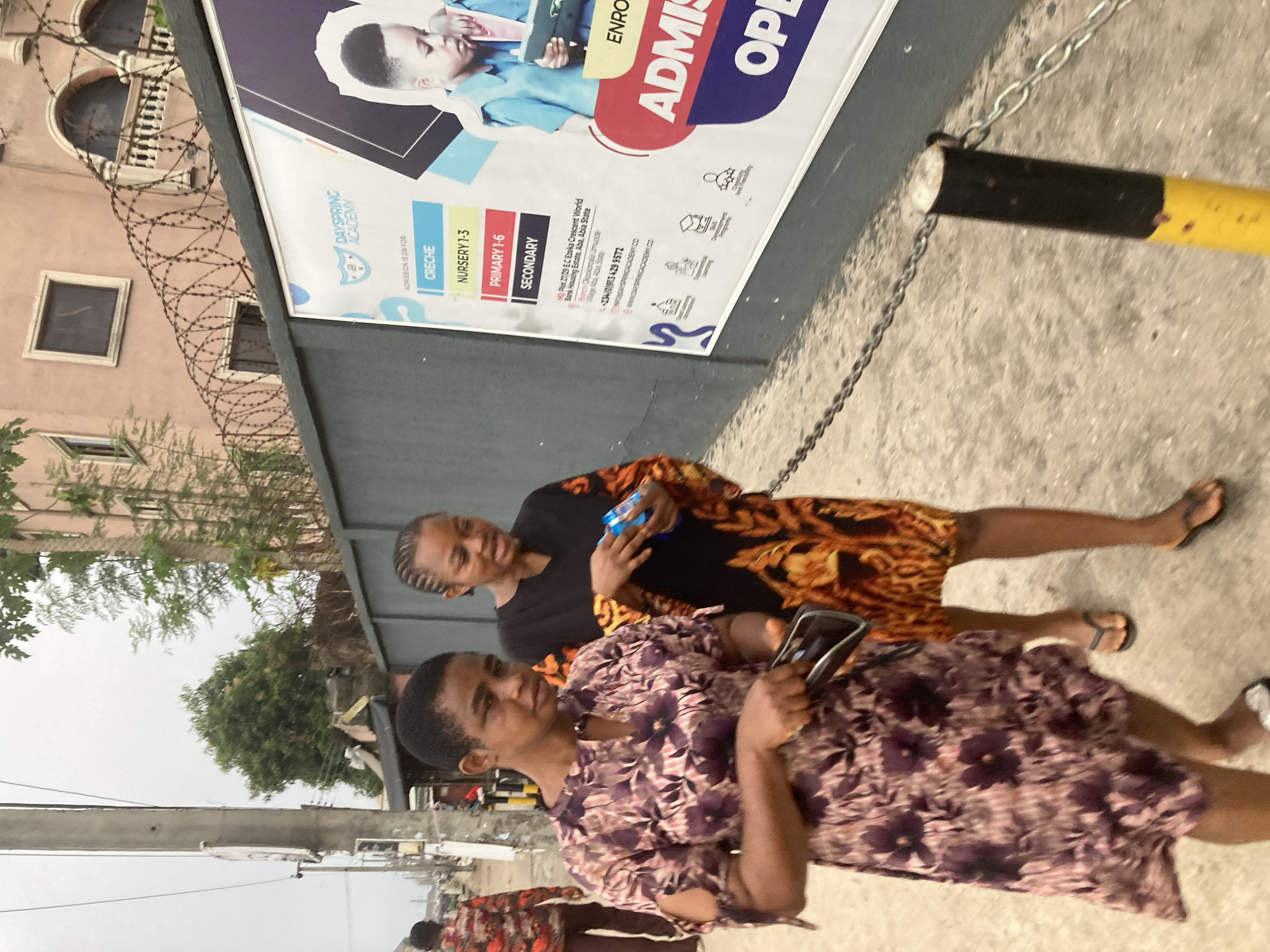 The image shows two women walking on a sidewalk in front of a building in Aba, Nigeria. The building appears to be Dayspring Academy, as indicated by the large sign with "DAYSPRING ACADEMY" printed on it. The sign also lists the school's programs, including Creche, Nursery 1-3, Primary 1-6, and Secondary. Additionally, the address is listed as Plot 27/29 EC Ezeka Crescent World Bank Housing Estate, Aba, Abia State.
One of the women is wearing a purple floral dress, holding a small black object. The other woman is wearing a black and orange patterned dress and appears to be holding a small blue container. The building behind the sign features barred windows and barbed wire fencing. The background shows a street with trees and buildings.