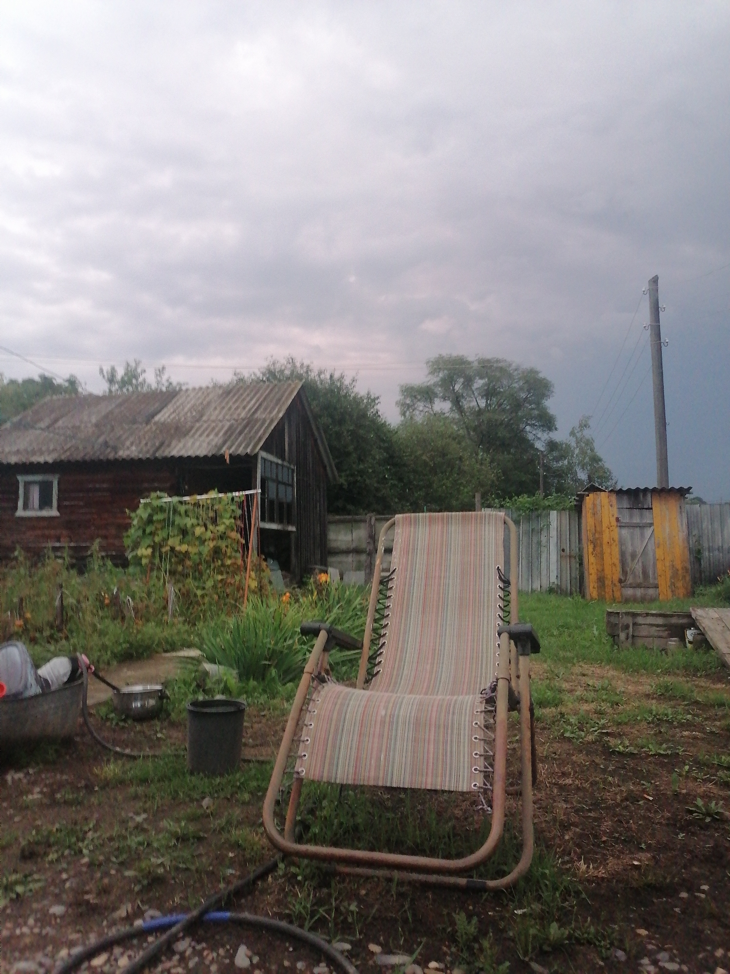 The image depicts an outdoor scene in what appears to be a rural or semi-rural setting, possibly a backyard or garden area. The foreground is dominated by a striped lounge chair, positioned at a slight angle. To the left of the chair, a large, gray, open-top container is visible, containing some objects, and a metal pot is placed nearby. A black plastic bucket sits on the ground behind these items. A dark hose is coiled on the ground in the lower part of the frame.
In the mid-ground, a wooden structure, likely a shed or small house, is visible with a corrugated roof and a single window. Green foliage and plants are growing around and in front of this structure, with some yellow flowers also present. Behind the wooden building, a wooden fence runs horizontally across the image. Further to the right, a small, yellow-painted wooden structure with a sloped roof and a door is situated near the fence.
The background consists of dense trees and vegetation. The sky is overcast with a grey, cloudy appearance. A wooden utility pole with wires is visible on the right side of the image, indicating the presence of electricity infrastructure. The overall atmosphere suggests a cloudy day, possibly before or after rain. The location is identified as Novopokrovka, Russia. There are no people or animals visible in the scene.