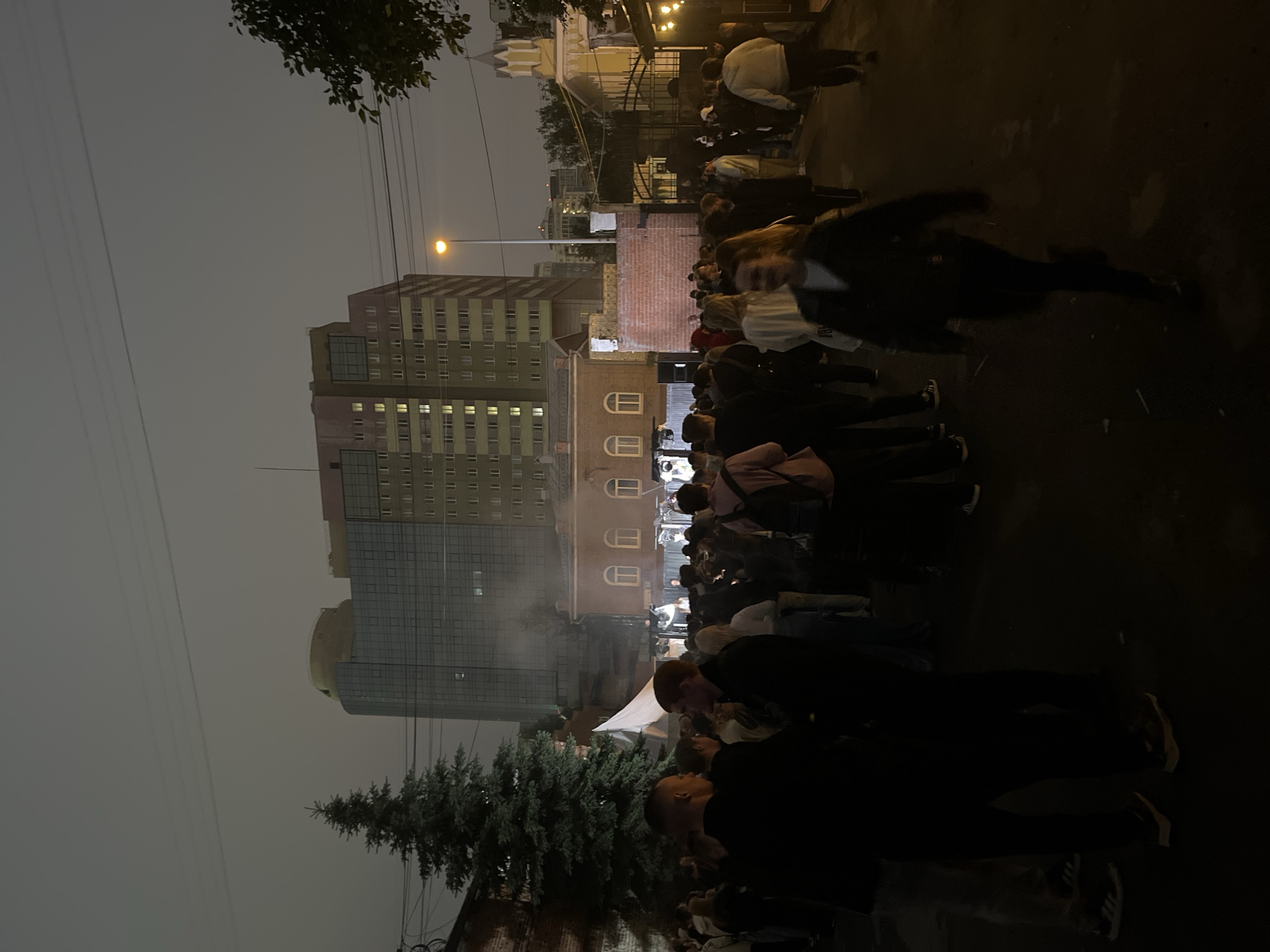 The image depicts an outdoor scene at dusk or night in a city. A crowd of people is gathered in what appears to be a street or open area. Several buildings are visible, including a tall, modern building with many windows in the background, and a lower, older building with arched windows in the middle ground. A large evergreen tree stands to the left of the frame. The lighting suggests artificial illumination from streetlights and possibly lights from within the buildings. Some individuals are walking, while others appear to be standing and socializing. The overall atmosphere is that of a gathering or event.