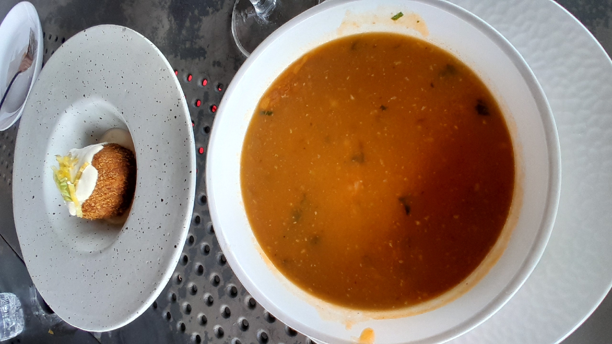 Two distinct dishes are presented on a perforated dark gray metal table in Pampatar, Venezuela. In the foreground, a white, round bowl contains an orange-red soup, characterized by a thick consistency and visible particulates, including small green specks, indicating herbs or vegetables. This bowl rests on a larger white plate with a subtle textured pattern. In the mid-ground, a shallow, wide-rimmed gray ceramic bowl, speckled with darker dots, holds a single, deep-fried, golden-brown spherical item. This item is topped with a white creamy sauce and garnished with yellow lemon zest and thin green leaf slivers. To the upper left, a portion of a clear water bottle is visible. To the upper right, a small white rectangular plate with a silver fork is present. A glass stem is partially visible along the right edge. Red light reflections are intermittently visible through the table's perforations. No individuals are present in the frame.