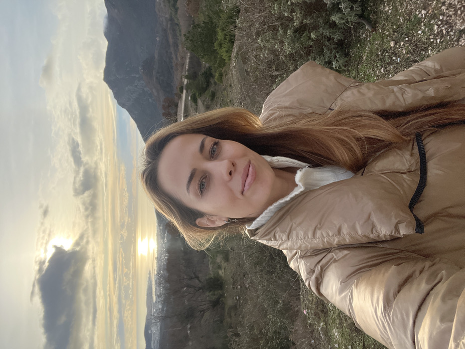 A woman poses for a selfie outdoors. She has fair skin, light brown hair, and wears a beige puffer jacket over a white top. Behind her, a mountainous landscape unfolds under a sky lit by the setting sun. The sun's rays peek through clouds, creating a golden hue over the scene. Some vegetation surrounds the immediate area. A town or city is visible in the distance.