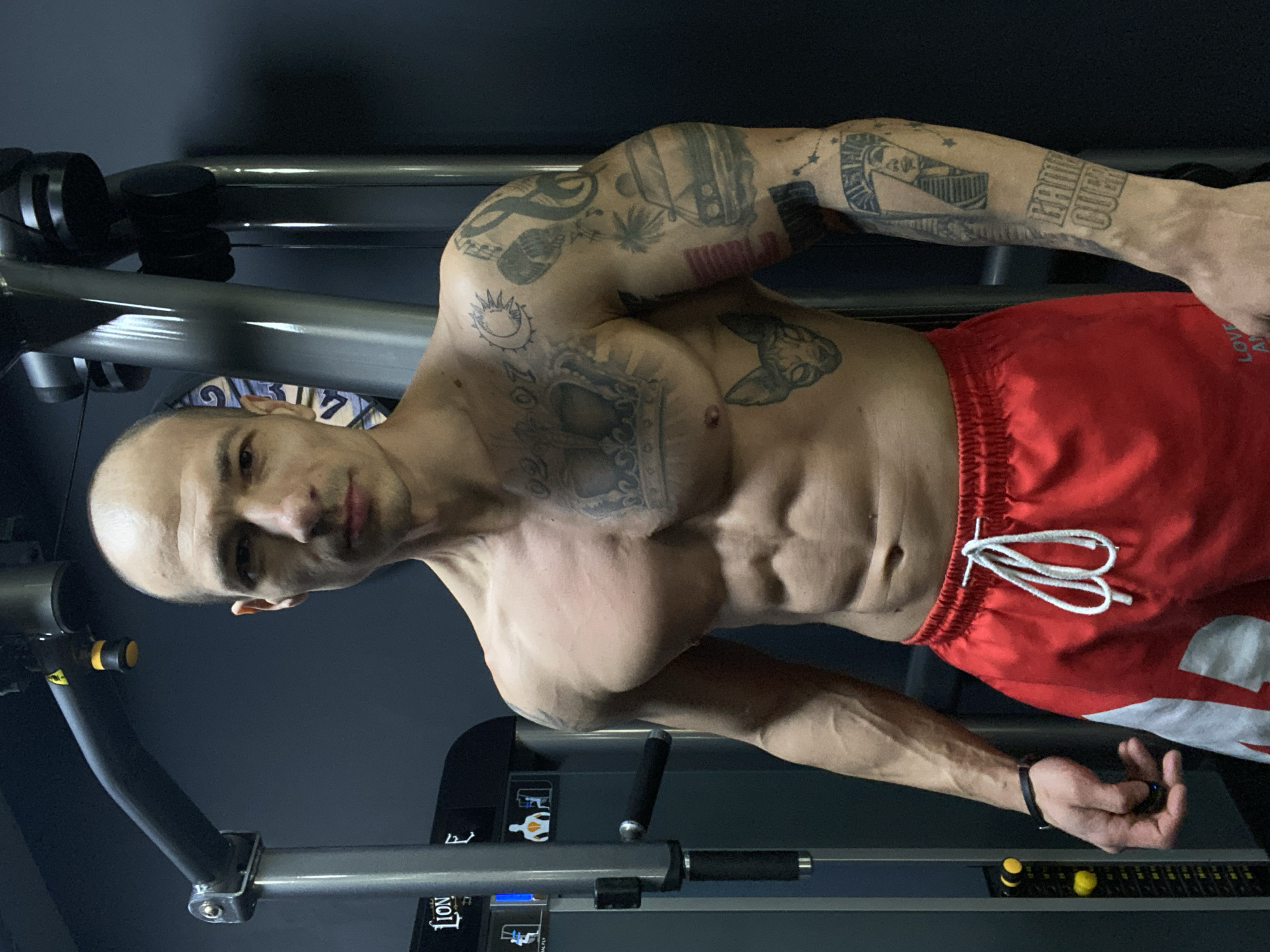 A muscular, bald man stands facing the camera in what appears to be a gym. He is shirtless, revealing a defined physique and extensive tattoos on his upper body and arms. He wears red shorts with a white drawstring. The background includes gym equipment with metallic and black elements. A partial view of a city is not visible. This event could not be identified to have happened in Pleven, Bulgaria.