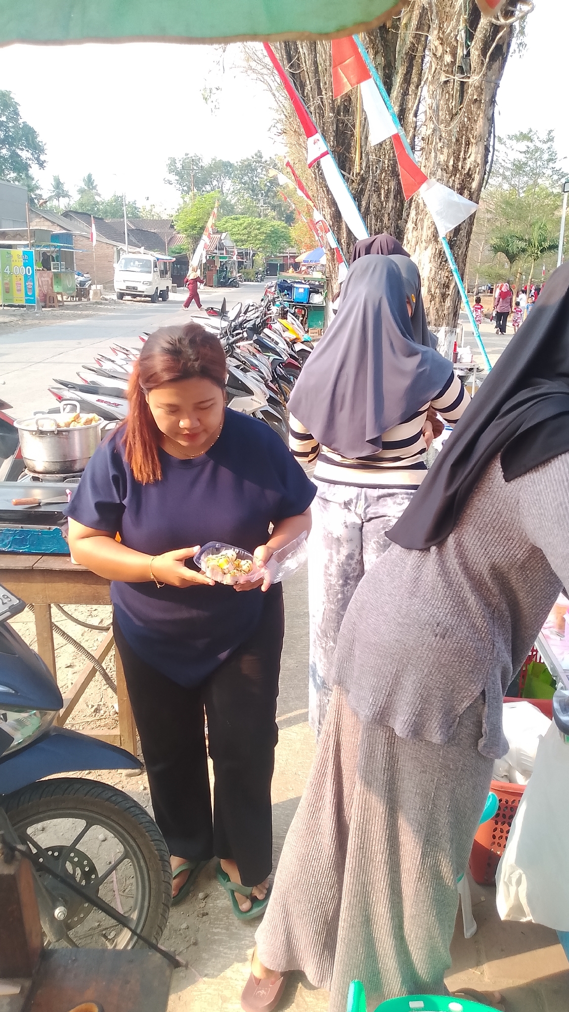 The image depicts an outdoor scene at a street market in Karanglangu, Indonesia, on a bright, sunny day. A woman with reddish-brown hair tied in a ponytail is standing in the foreground, holding a clear plastic container filled with food items. She is wearing a dark blue t-shirt and black pants, with green flip-flops on her feet. To her right, a person wearing a grey hijab and a ribbed grey maxi dress is partially visible, also with their feet shod in brown flat shoes. Further back, another person in a dark hijab and a striped long-sleeved shirt is seen with patterned pants.
The background reveals a street lined with parked motorcycles. Red and white Indonesian flags are strung up, attached to poles and trees. In the distance, there are buildings, including shops, and a white van is visible on the road. The ground appears to be a mix of dirt and paved surfaces. A food stall is set up with a pot and other cooking utensils. The overall atmosphere suggests a local community gathering or marketplace.
