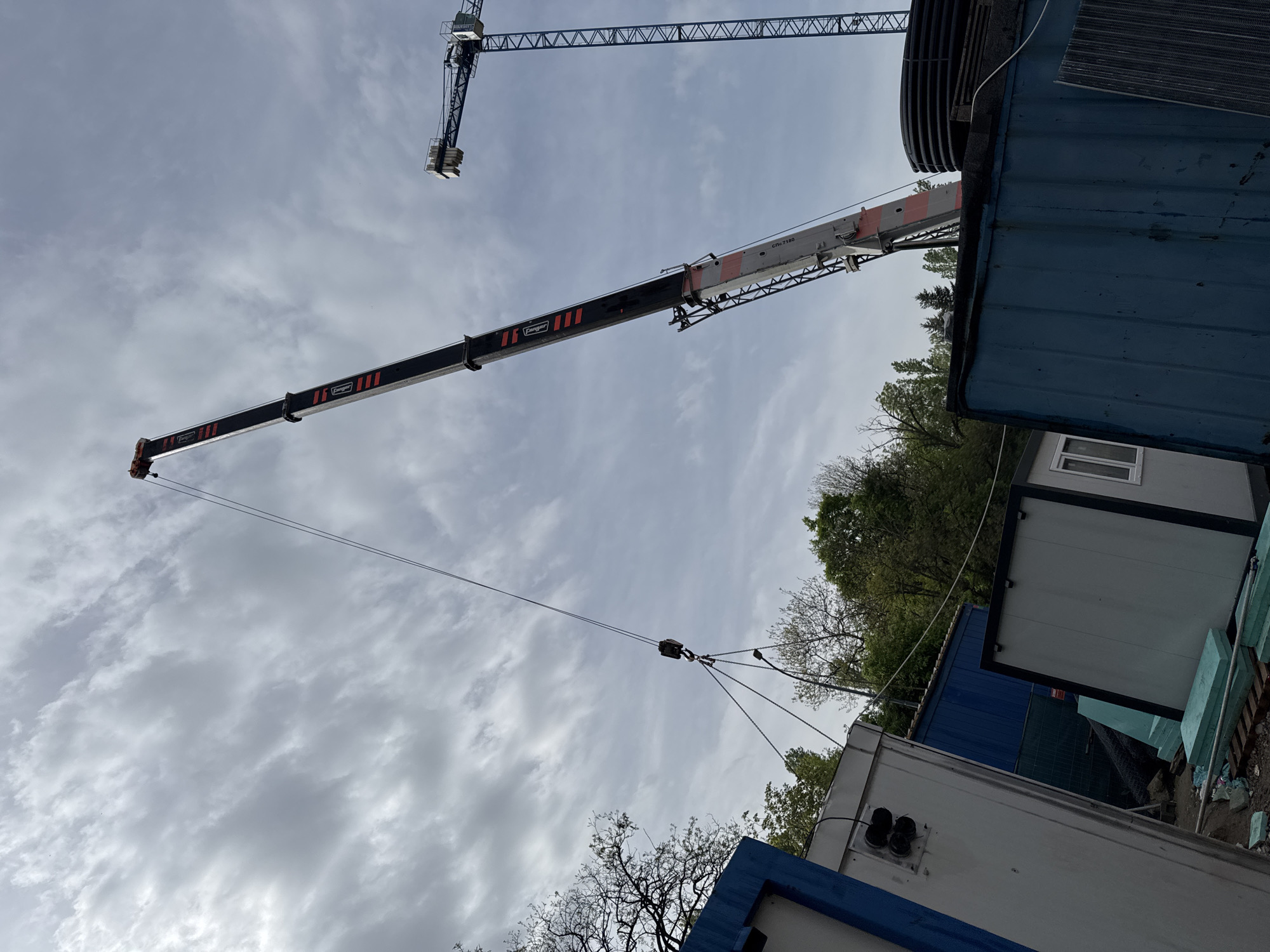 The image shows a construction site, possibly in the outskirts of a city. Several portable modular buildings are arranged next to each other. In the background, there are trees and foliage suggesting a natural setting nearby. Two construction cranes are visible in the sky, with one crane's boom extended and rigged with cables and a hoist. The sky is overcast with clouds. There are no people visible in the image. The ground is partially obscured by the modular buildings.