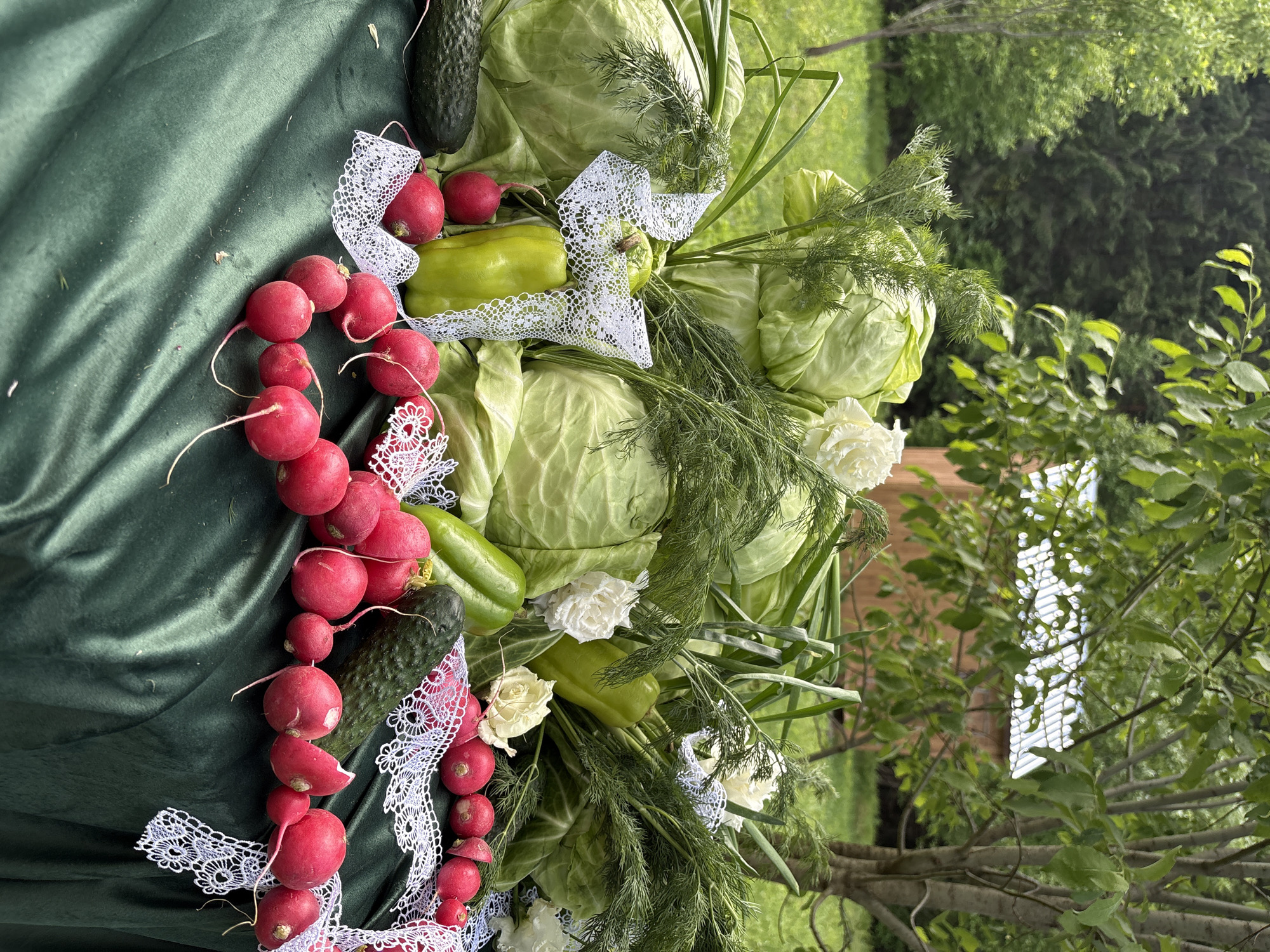 The image shows a still life arrangement of vegetables and flowers. The vegetables include cabbages, radishes, green peppers, and cucumbers, arranged with dill and white flowers, possibly roses. A piece of white lace adorns the vegetables. The arrangement is placed on a green velvet-like cloth. The background includes outdoor scenery with green foliage and trees. The location is stated as Sibir', Russia, which may be a clue to the arrangement's purpose.