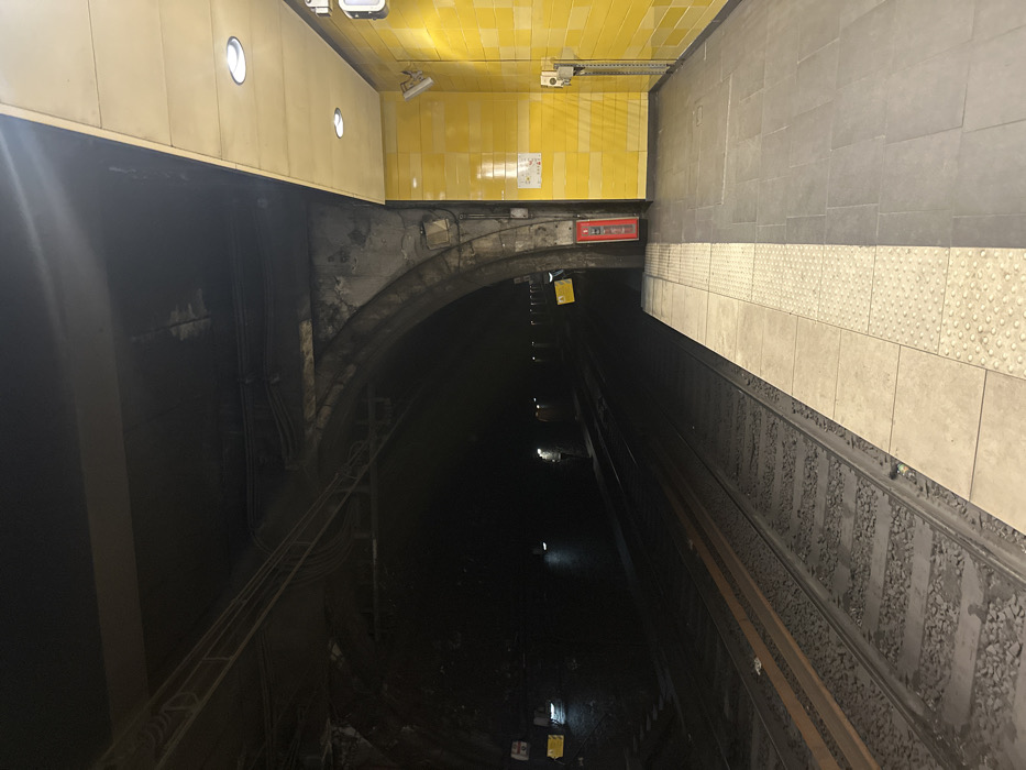 This image depicts an underground train station platform and associated tunnel, located in Vincennes, France. The scene is devoid of human presence or activity.
On the left side of the frame, two parallel railway tracks, supported by ballast, recede into a dark, concrete-arched tunnel. The tunnel interior is dimly lit by scattered overhead lights, revealing multiple sets of cables and conduits running along the arch structure and ceiling. Several rectangular yellow warning signs with dark text/symbols are affixed to the tunnel wall at intervals.
The platform, occupying the right side of the image, is brightly illuminated by multiple recessed circular lights embedded in a flat, light-colored ceiling panel. Its floor is paved with large grey tiles, featuring a distinct light-colored tactile strip with raised circular bumps running along the edge adjacent to the tracks. The platform wall is faced with horizontally-arranged bright yellow ceramic tiles, displaying subtle variations in shade between horizontal bands. Essential objects mounted on this wall include a prominent red fire extinguisher contained within a red casing, positioned near the junction of the tunnel arch and platform wall. Further along the yellow tiling, a grey emergency communication box with a handset is mounted, alongside two adjustable wall-mounted light fixtures with white casings, directed towards the platform. A white rectangular information panel or map is also present on the yellow tiled wall above the fire extinguisher.