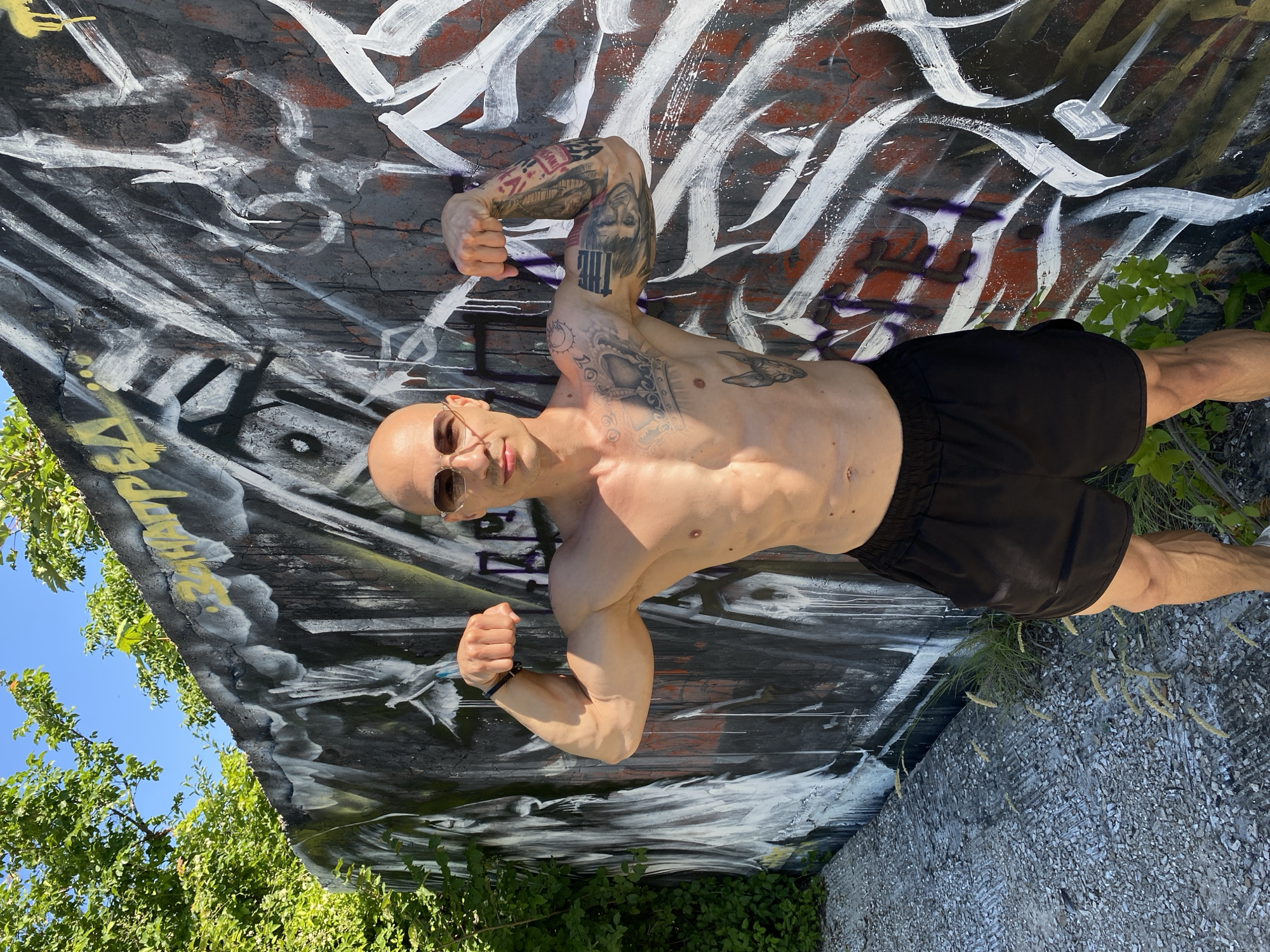 A bald, shirtless man with sunglasses and black shorts poses by a graffiti-covered wall in Pleven, Bulgaria. He is flexing his biceps, showcasing his muscular physique and tattoos on his arms and chest. The wall, part of an outdoor structure, is adorned with colorful graffiti and appears to be aged. Lush green foliage surrounds the area, and the ground is covered with gravel. The sky above is clear and blue, suggesting it is daytime with bright sunlight.